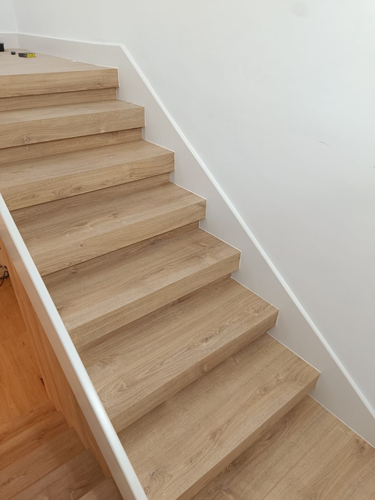 Una escalera con peldaños de madera clara, contrahuellas blancas y una pared de largueros blancos en el lado derecho.