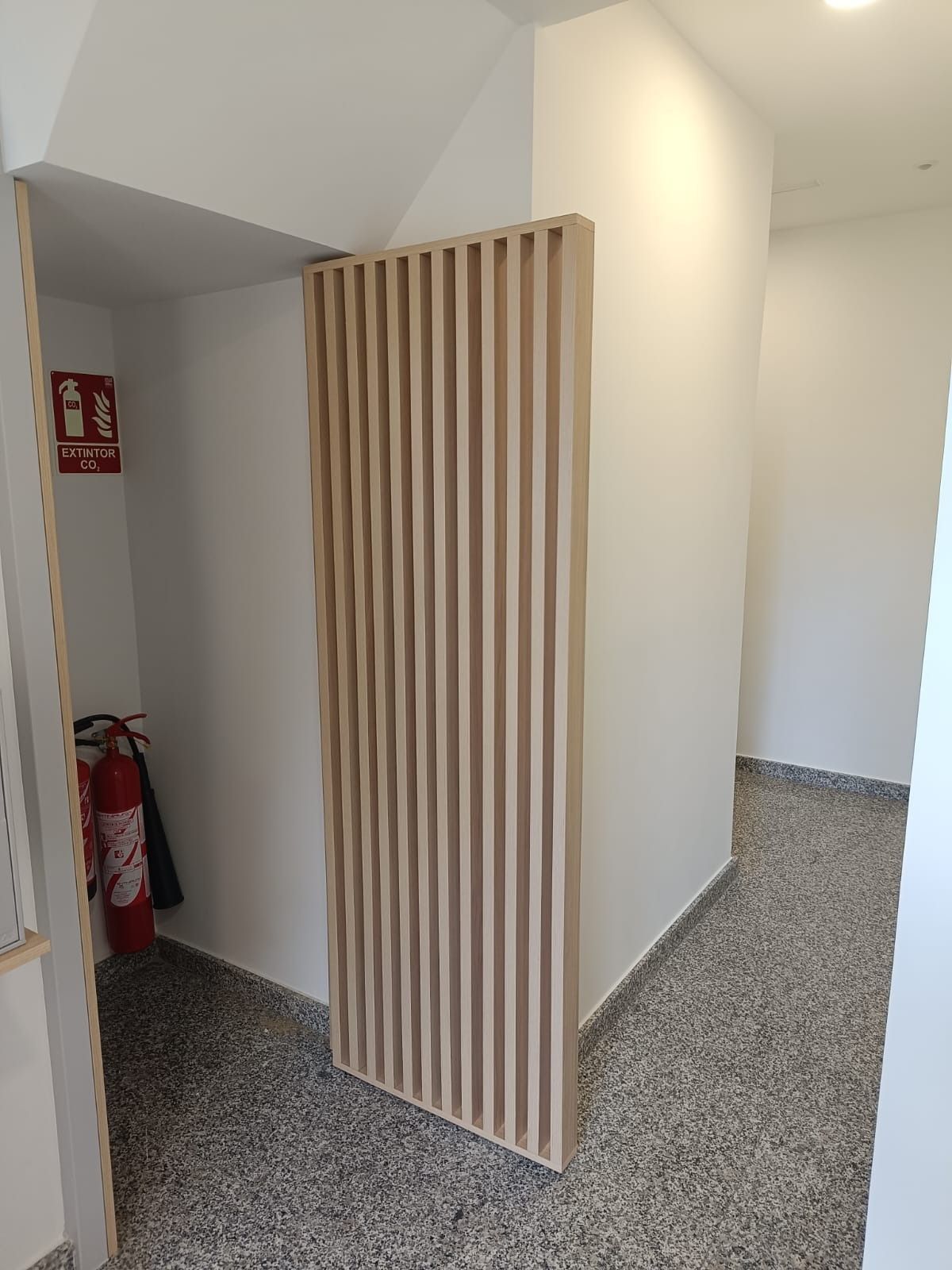 Una mampara de privacidad de listones de madera en un pasillo, con un extintor rojo en la pared cercana.