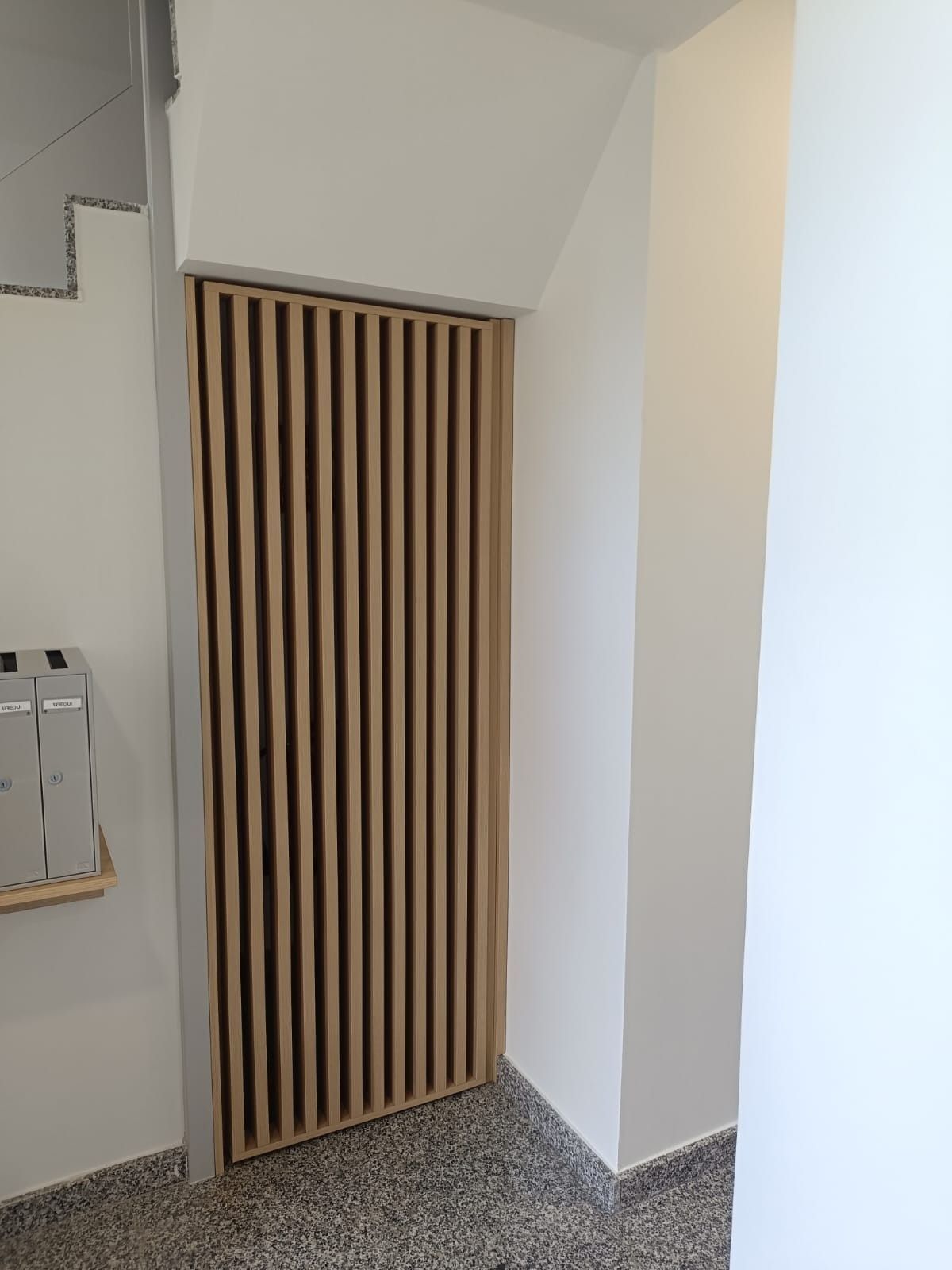 Una puerta de listones de madera instalada en un hueco de pared blanca sobre un suelo gris moteado.