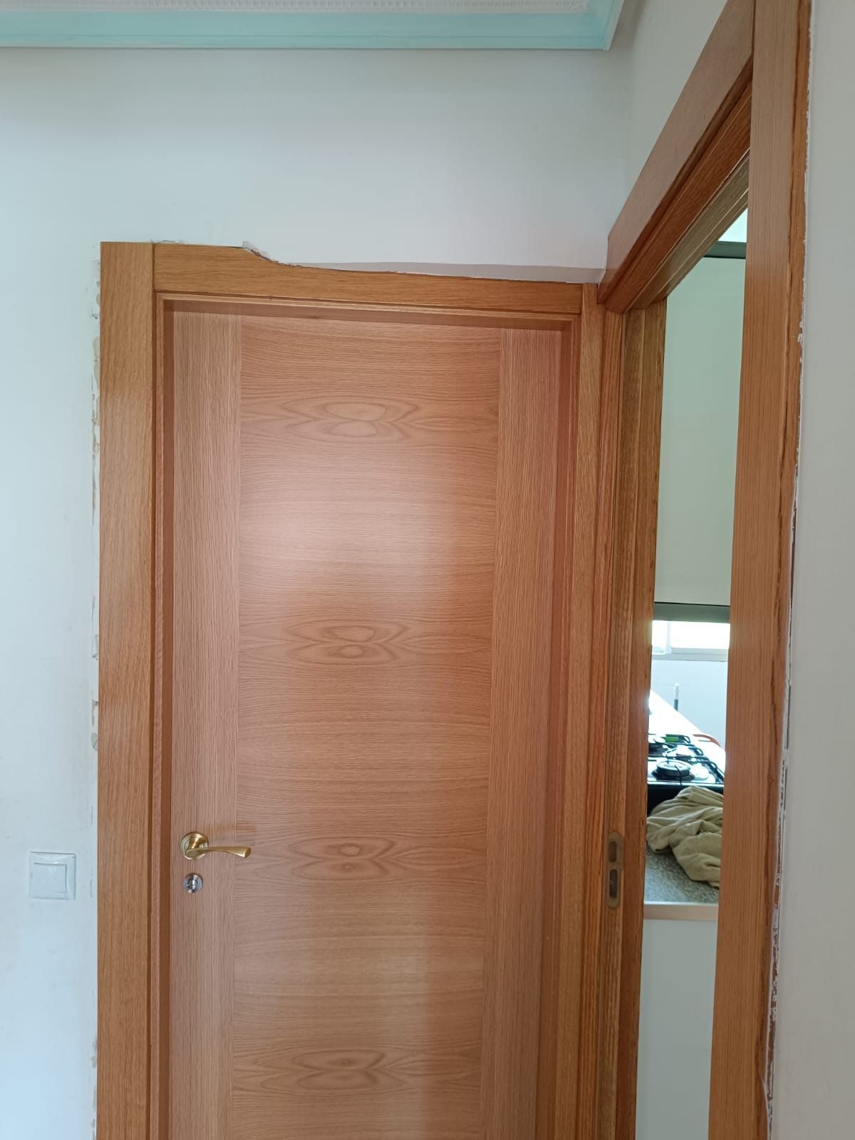 Una puerta de madera clara instalada en una pared blanca, con una puerta contigua que da a otra habitación.