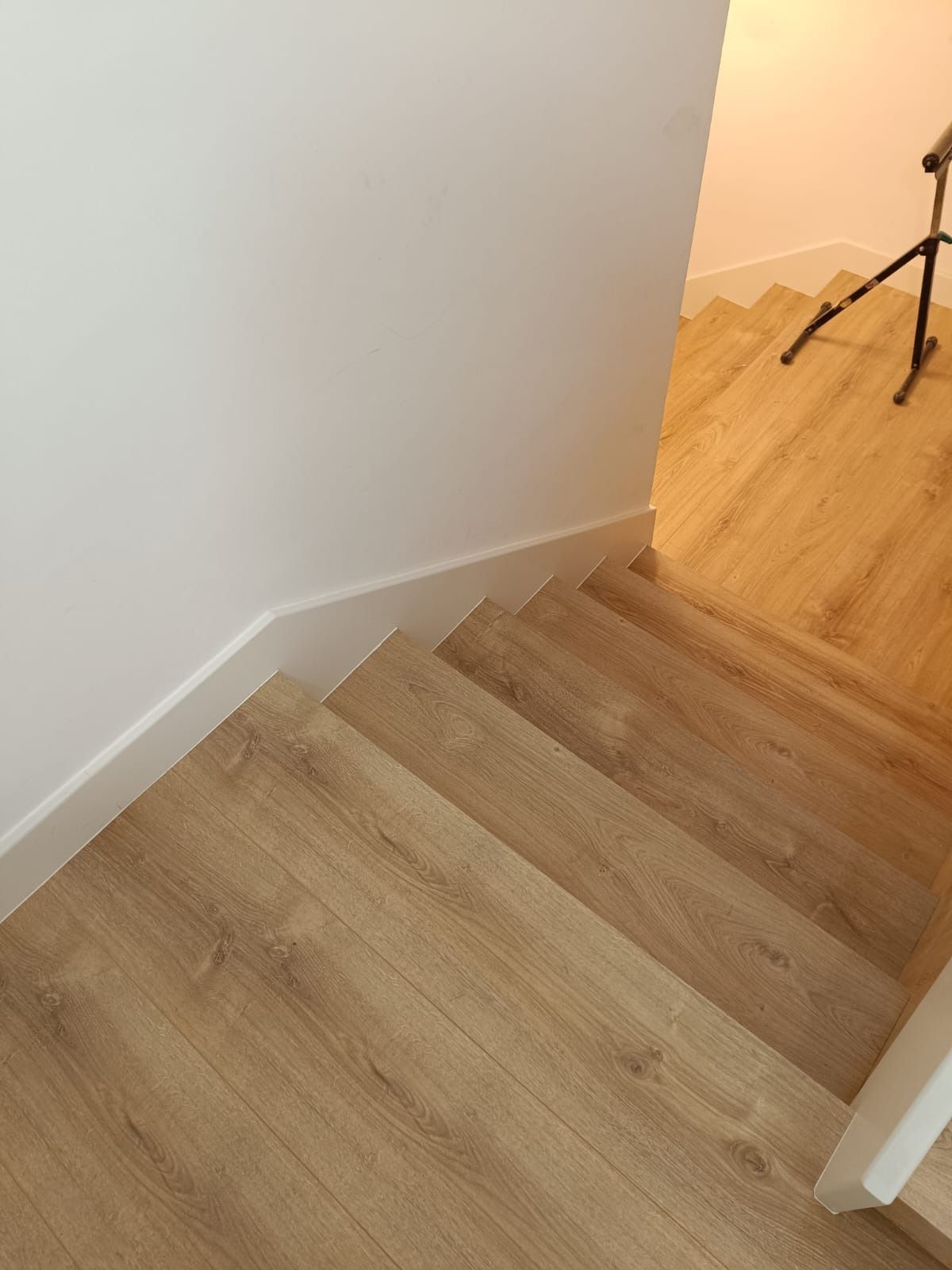 Vista desde arriba de una escalera de madera clara que desciende hacia un pasillo, con un trípode visible en la esquina.