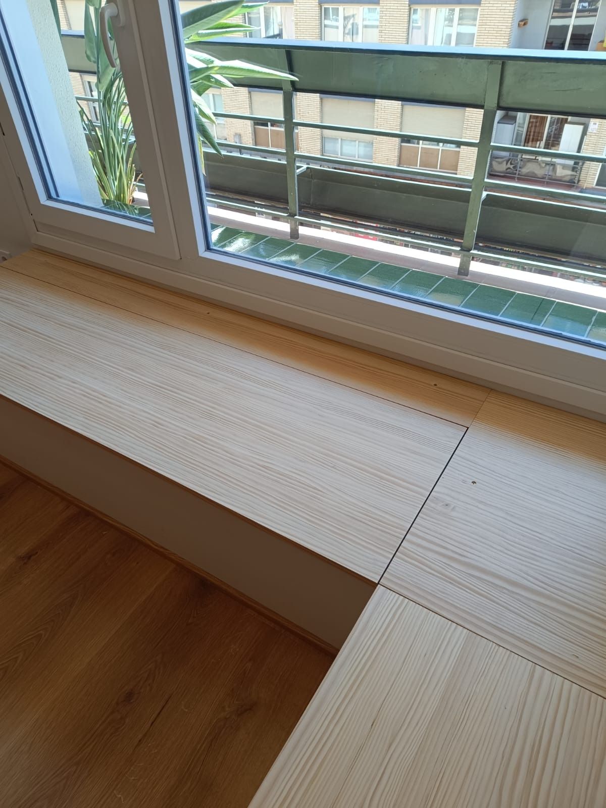 Un banco de madera de tono claro integrado en una esquina, situado junto a una ventana que da a un balcón.