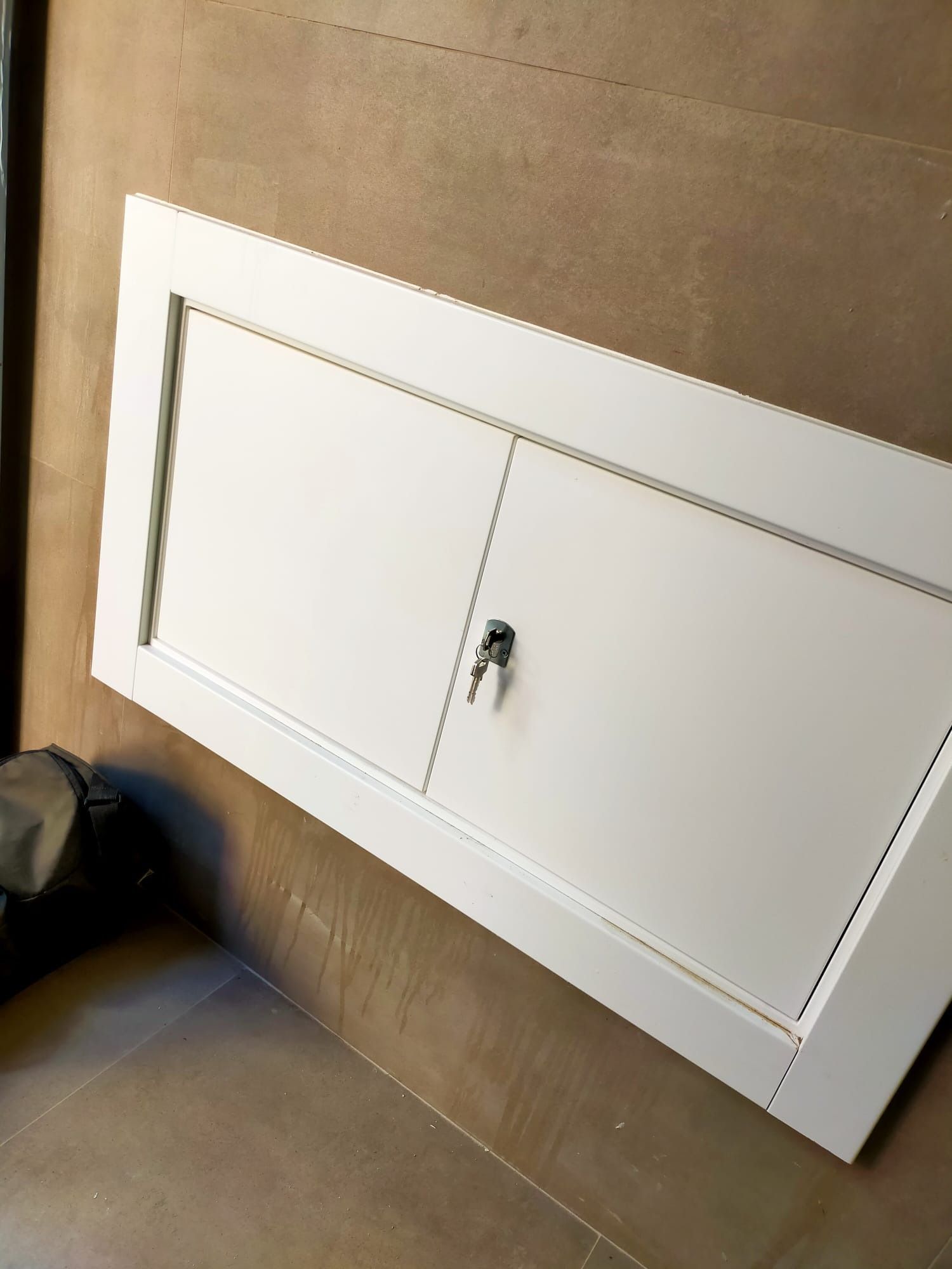 Un panel de acceso rectangular blanco con cerradura central, montado sobre una pared lisa de color beige.