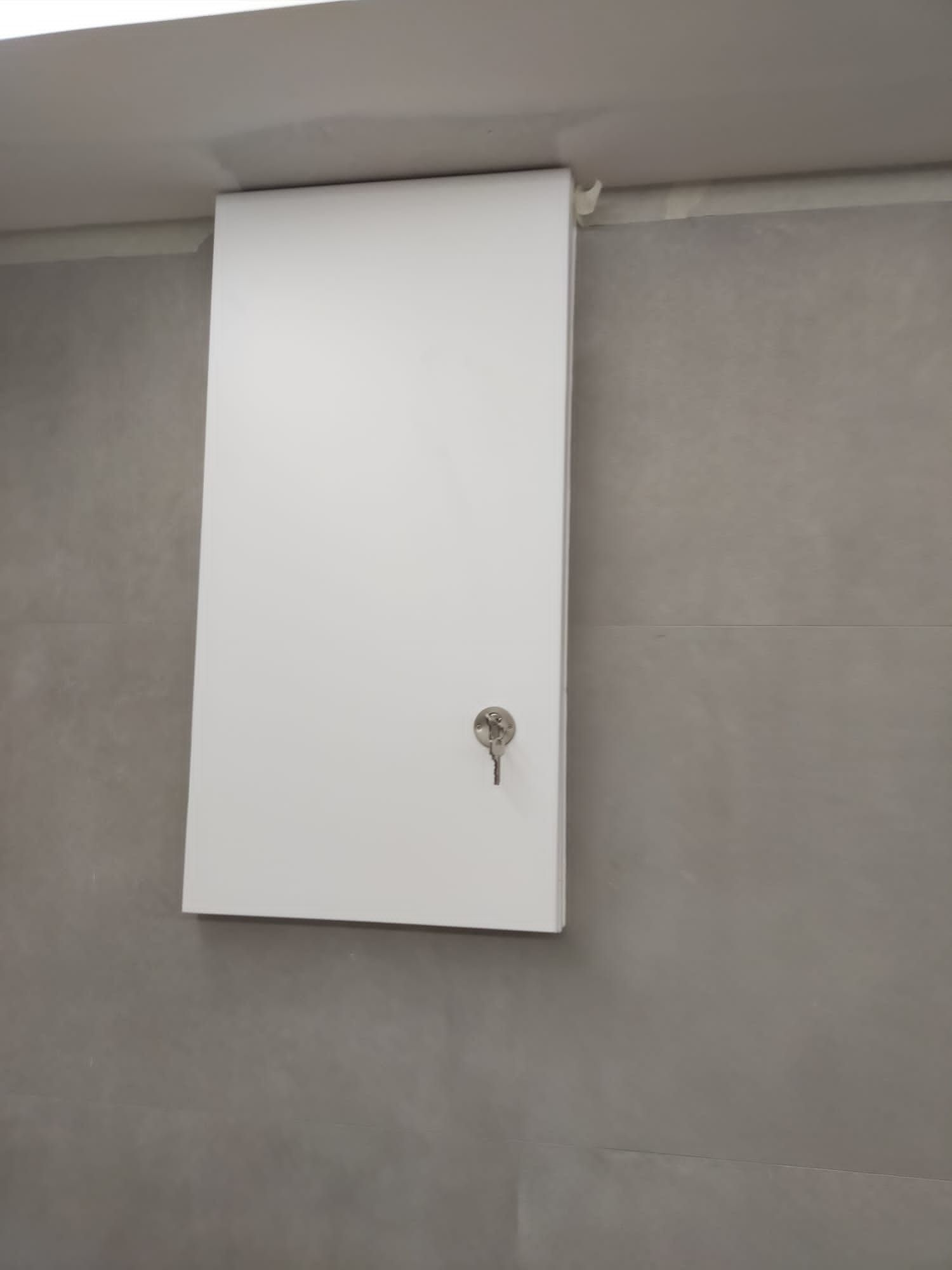 Un panel de acceso blanco para servicios públicos con cerradura, montado en una pared gris lisa cerca de un techo blanco.