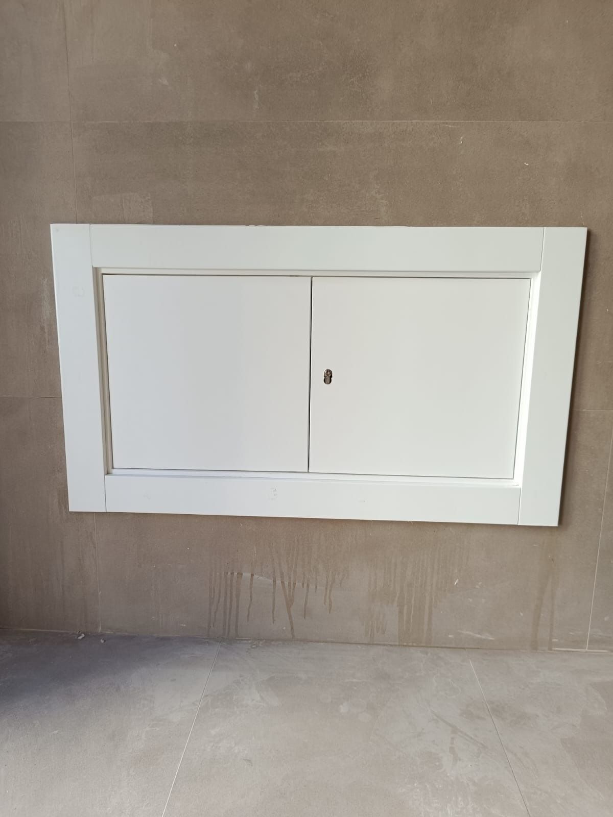 Un panel de acceso rectangular blanco con puertas dobles instalado a ras de una pared lisa de color marrón claro.
