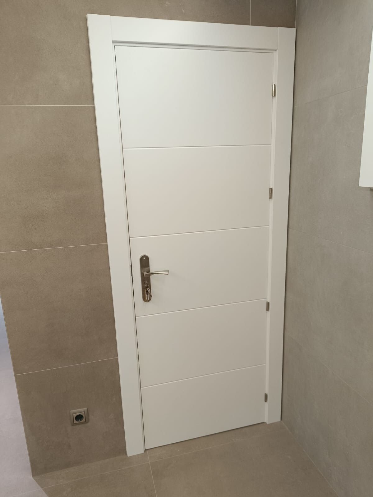 Una puerta interior blanca con paneles horizontales y tirador metálico, instalada en una pared alicatada.