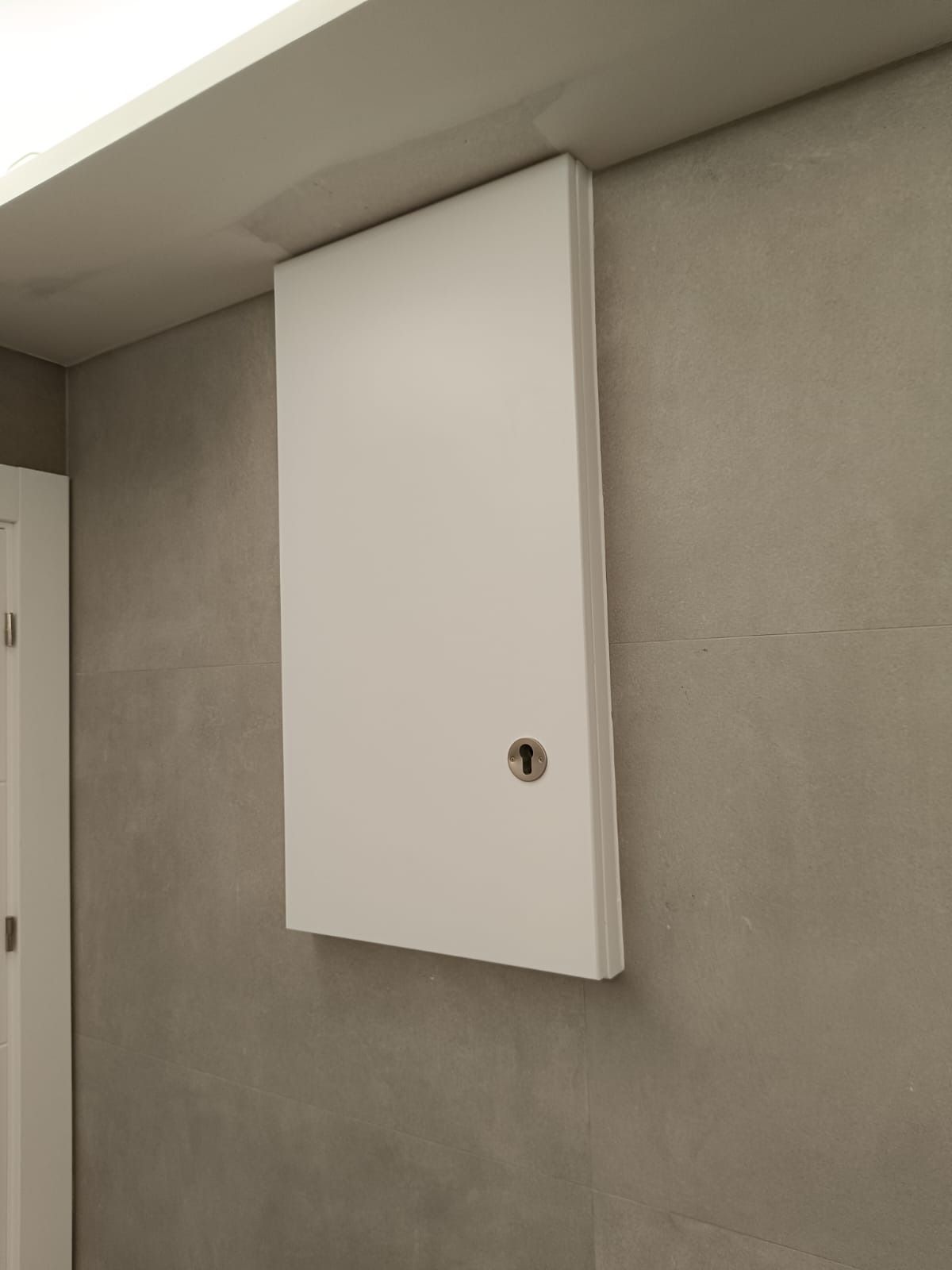 Un panel de acceso rectangular de color blanco con una cerradura, montado sobre una pared alicatada de color gris.