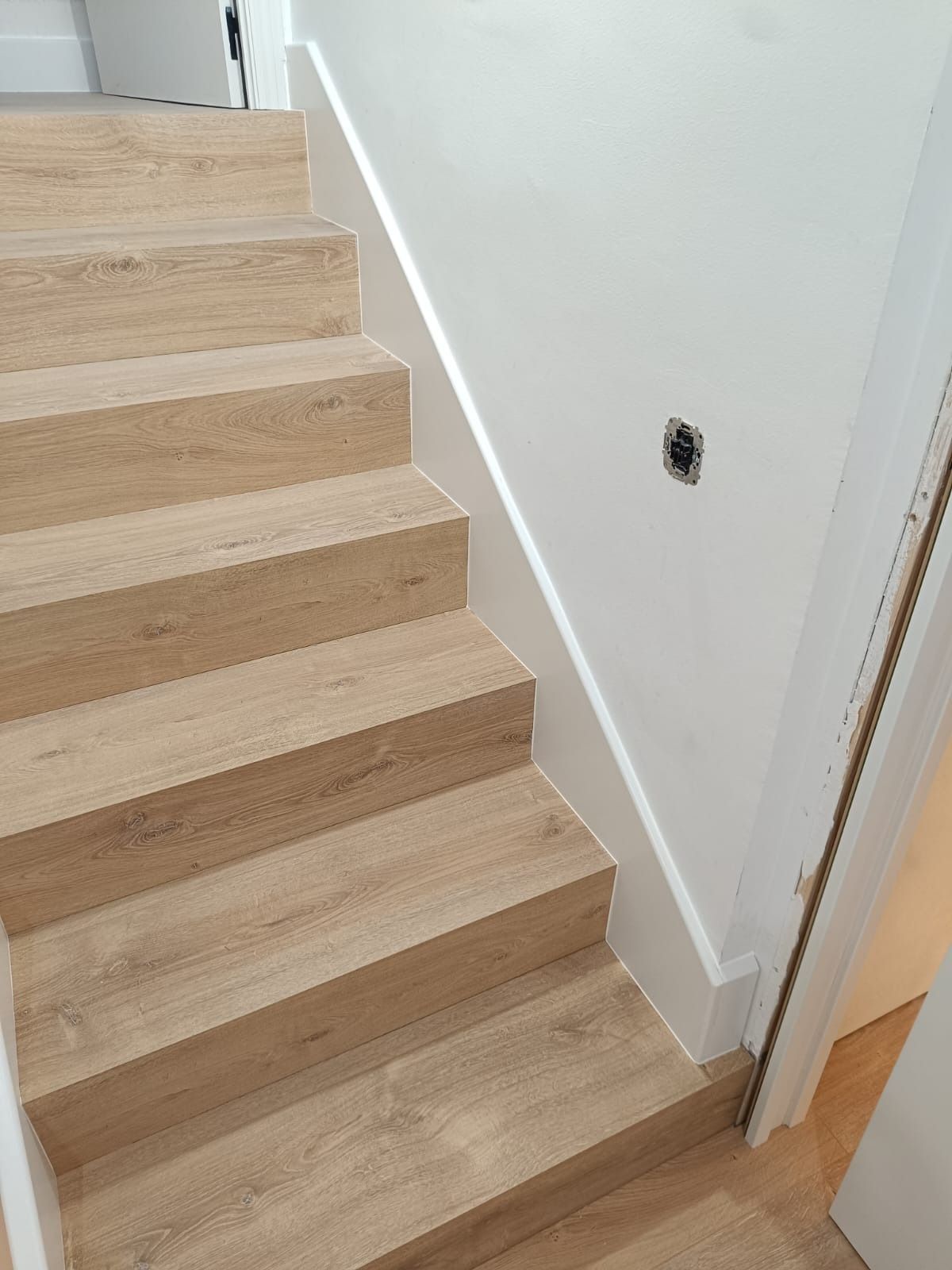 Escalera de madera clara que asciende junto a una pared blanca lisa con una caja de enchufes eléctricos a la vista.
