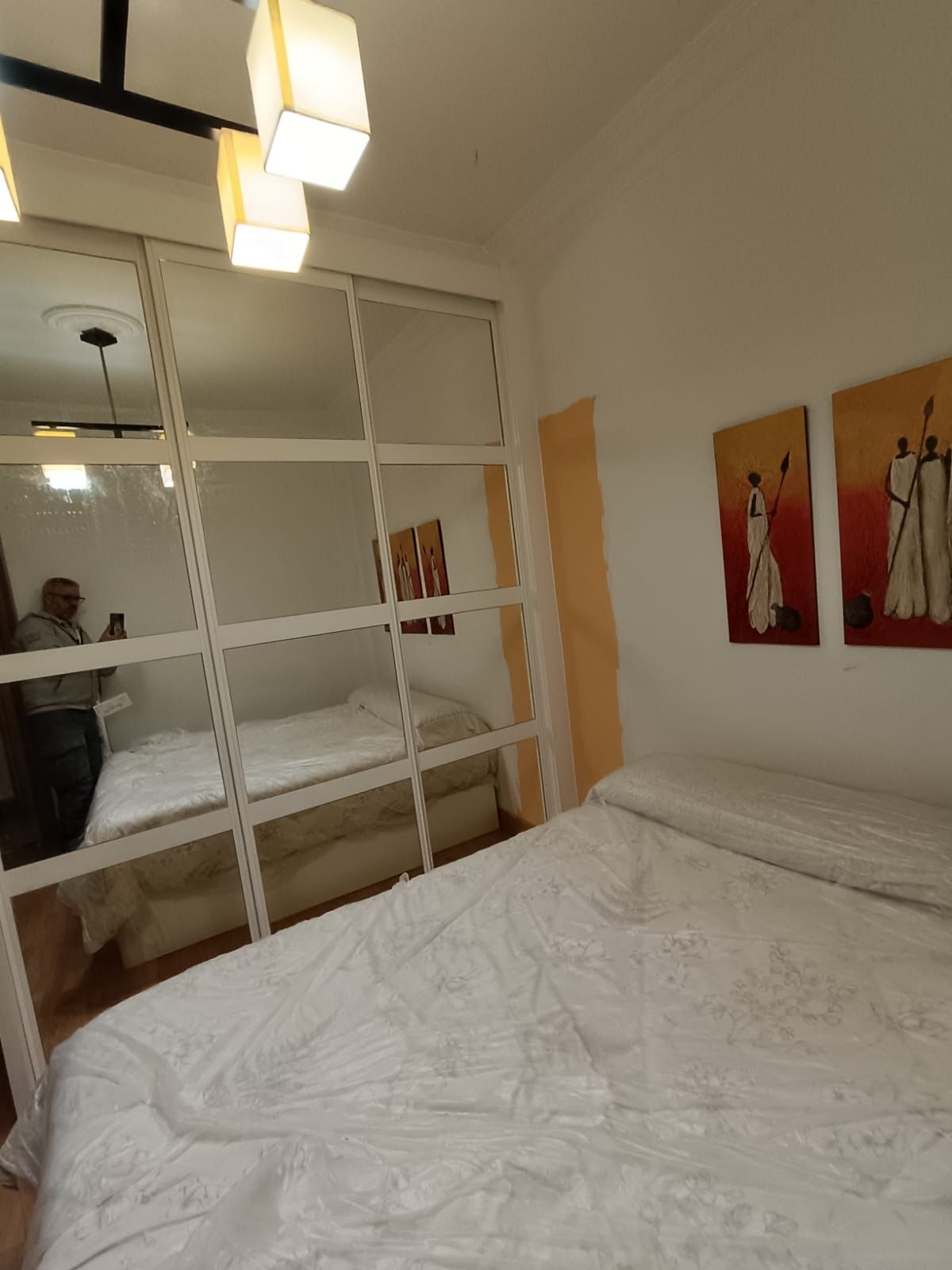 Un dormitorio con un gran armario con espejo que refleja una cama, y ​​dos cuadros enmarcados en la pared blanca contigua.