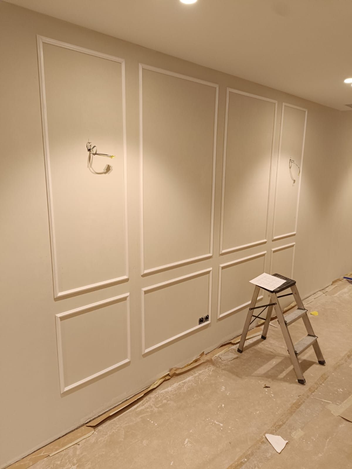 Una habitación en proceso de renovación con paredes de color claro, paneles decorativos con molduras y una escalera metálica.