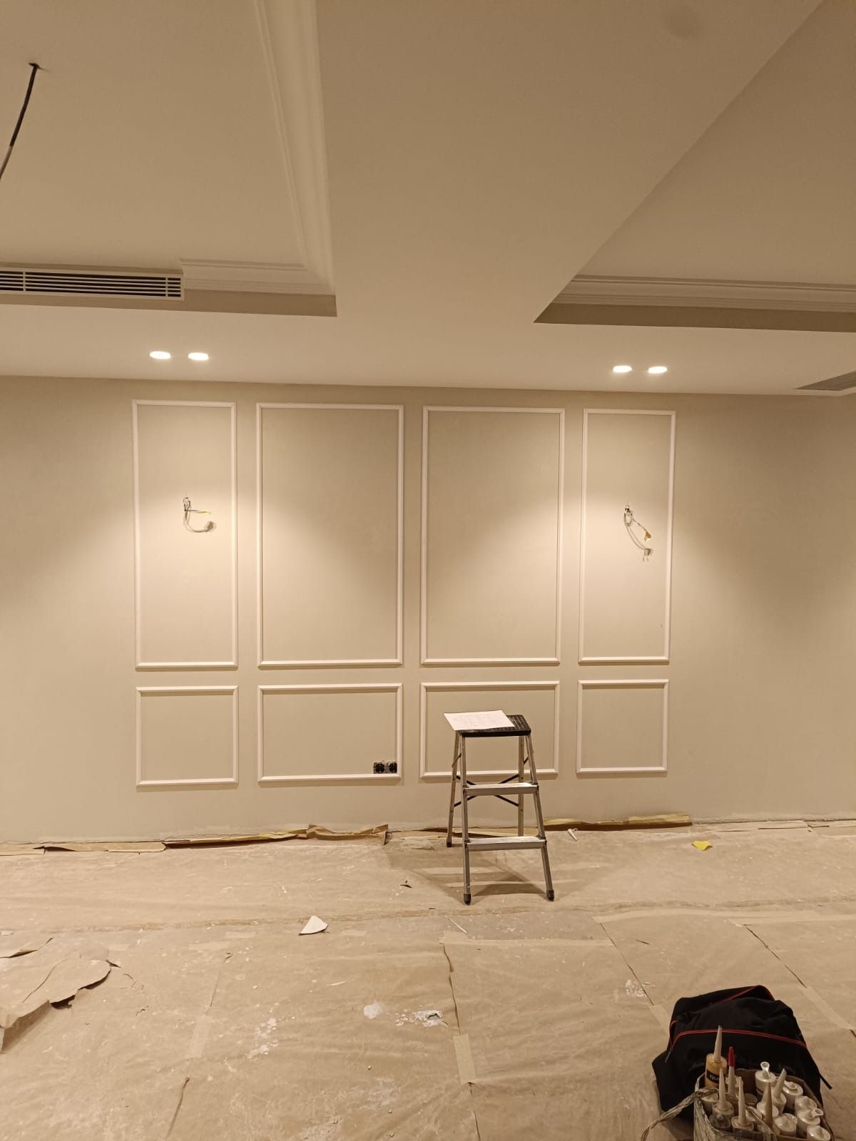 Una pared de color beige con molduras blancas simétricas bajo luces empotradas en el techo, con una escalera delante.