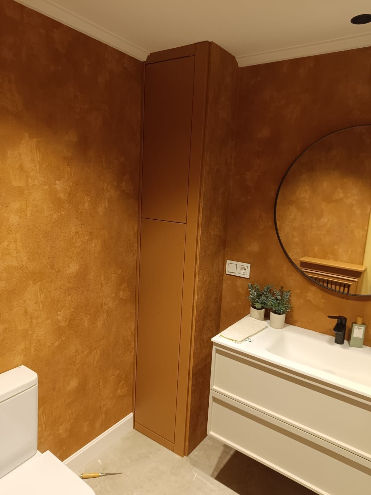 Un baño moderno con paredes de terracota cálida y texturizada, un armario alto a juego y un tocador blanco con lavabo.