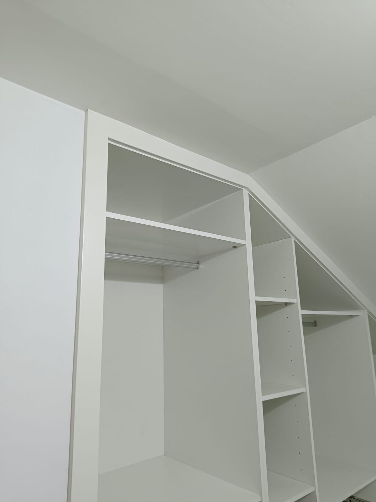 Un armario empotrado blanco con estantes abiertos y barras para colgar ropa, inclinado para adaptarse a un techo inclinado.