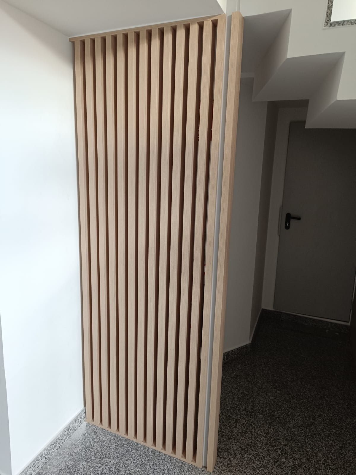 Un biombo vertical de listones de madera clara se alza en un pasillo con paredes blancas y suelo gris moteado.