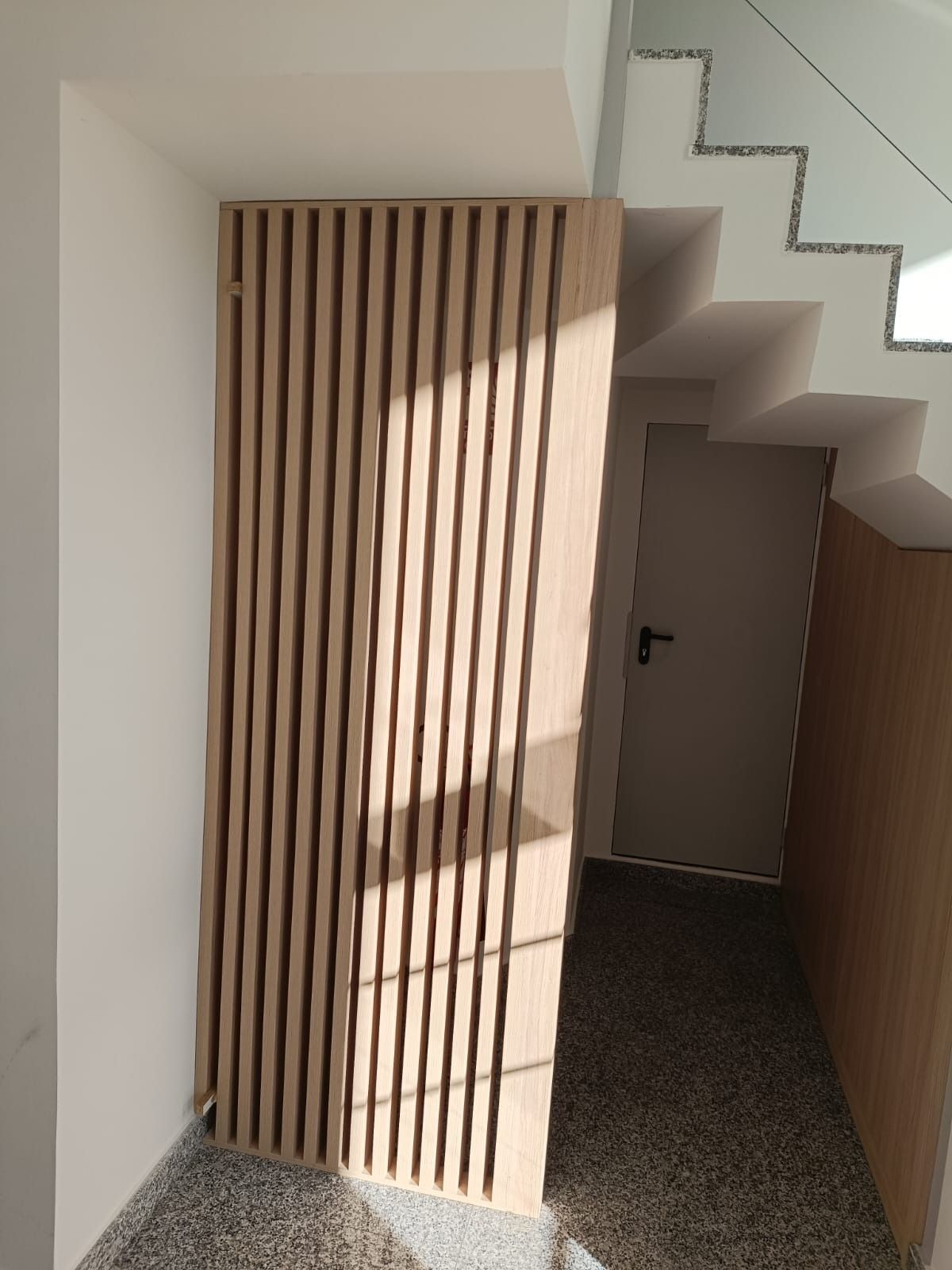 Un tabique vertical de listones de madera junto a una escalera y una puerta en un interior moderno con suelo moteado.