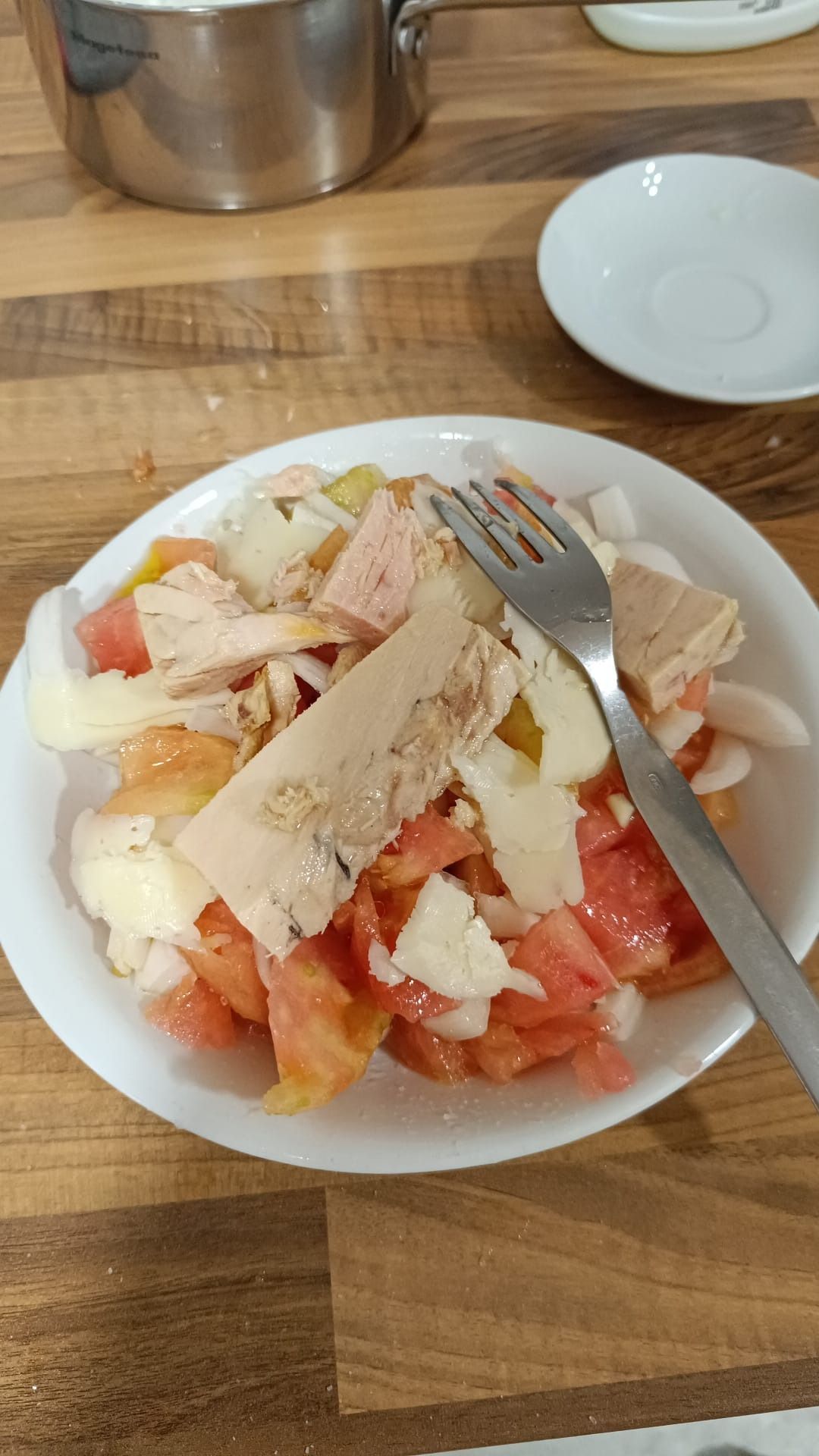 Una ensalada de tomates en rodajas, queso blanco y atún en un tazón blanco con un tenedor de plata sobre una mesa de madera.