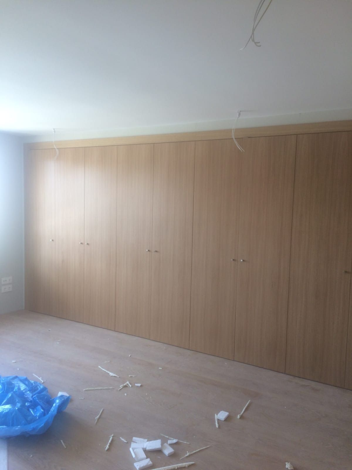 Una pared de armarios con acabado de madera clara en una habitación con suelo claro y escombros de construcción en el piso.