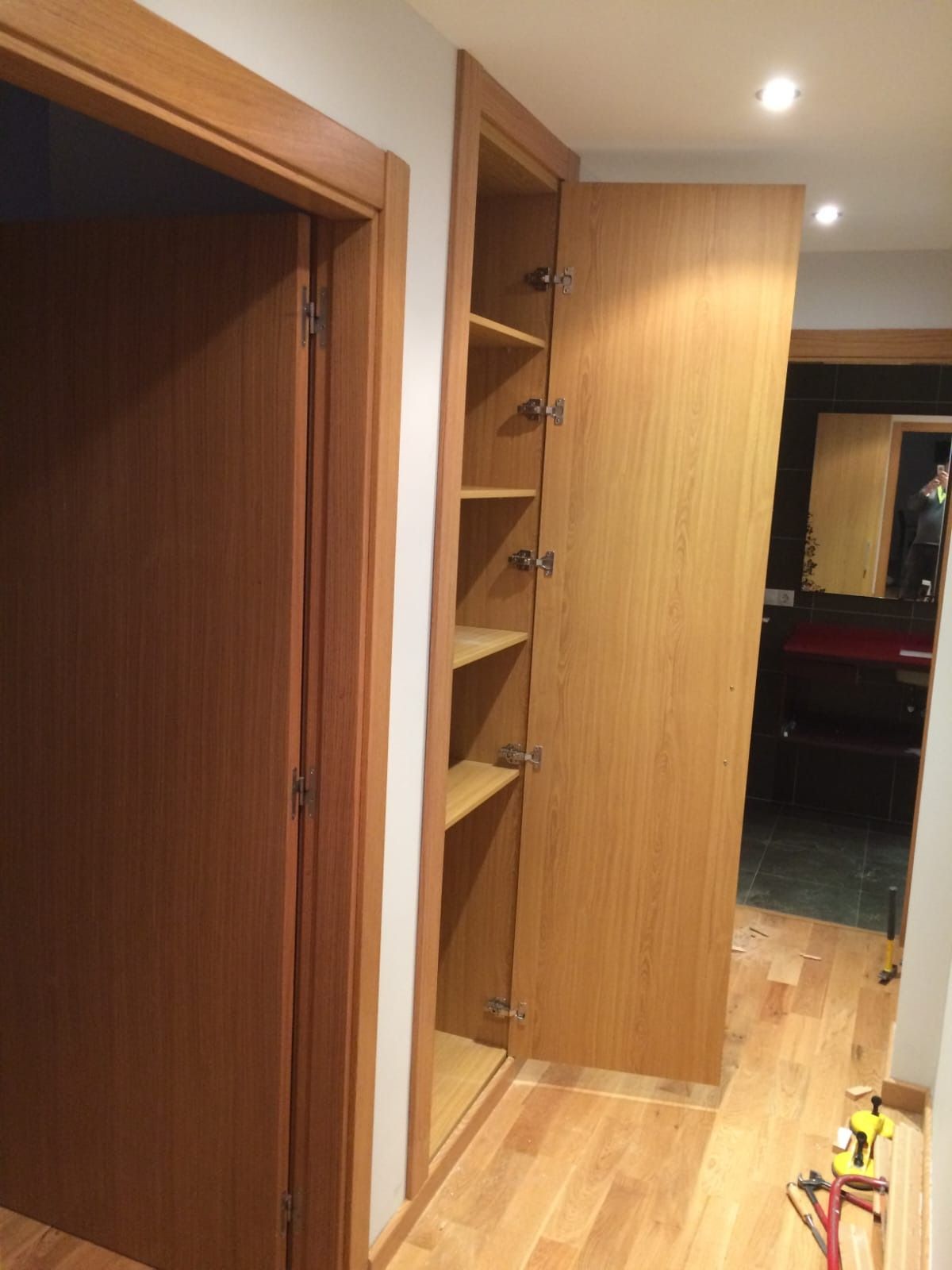 Una puerta de madera se encuentra junto a un armario alto y abierto con estantes en un pasillo con suelo de madera clara.