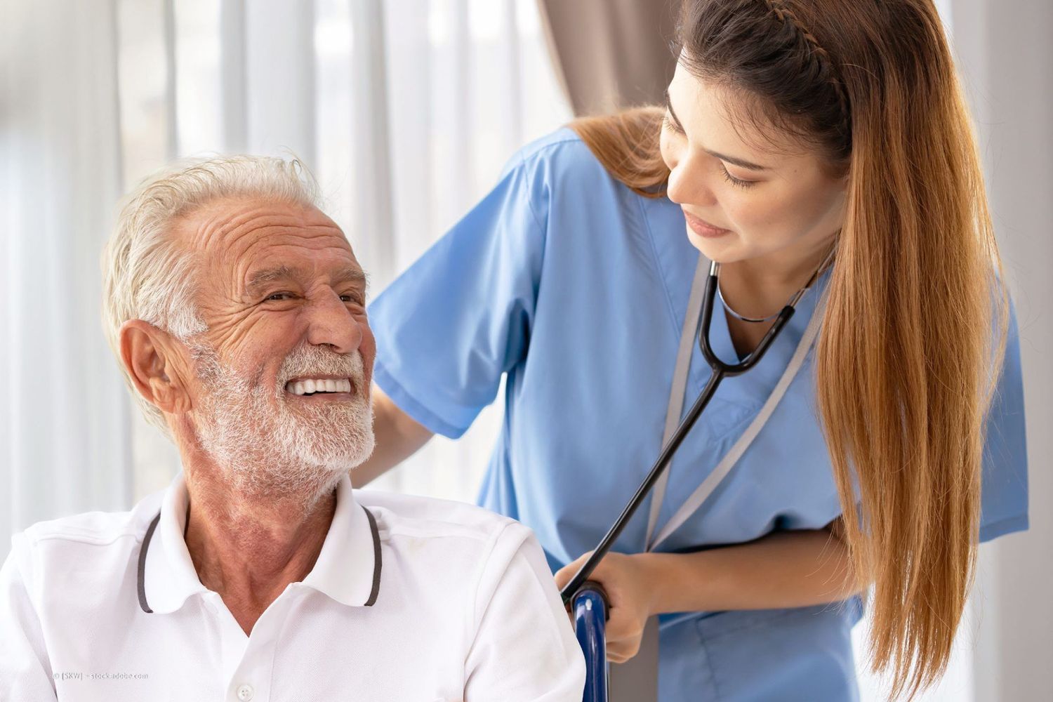 Krankenschwester mit Stethoskop hilft älterem Mann im Rollstuhl; beide lächeln im Innenbereich.