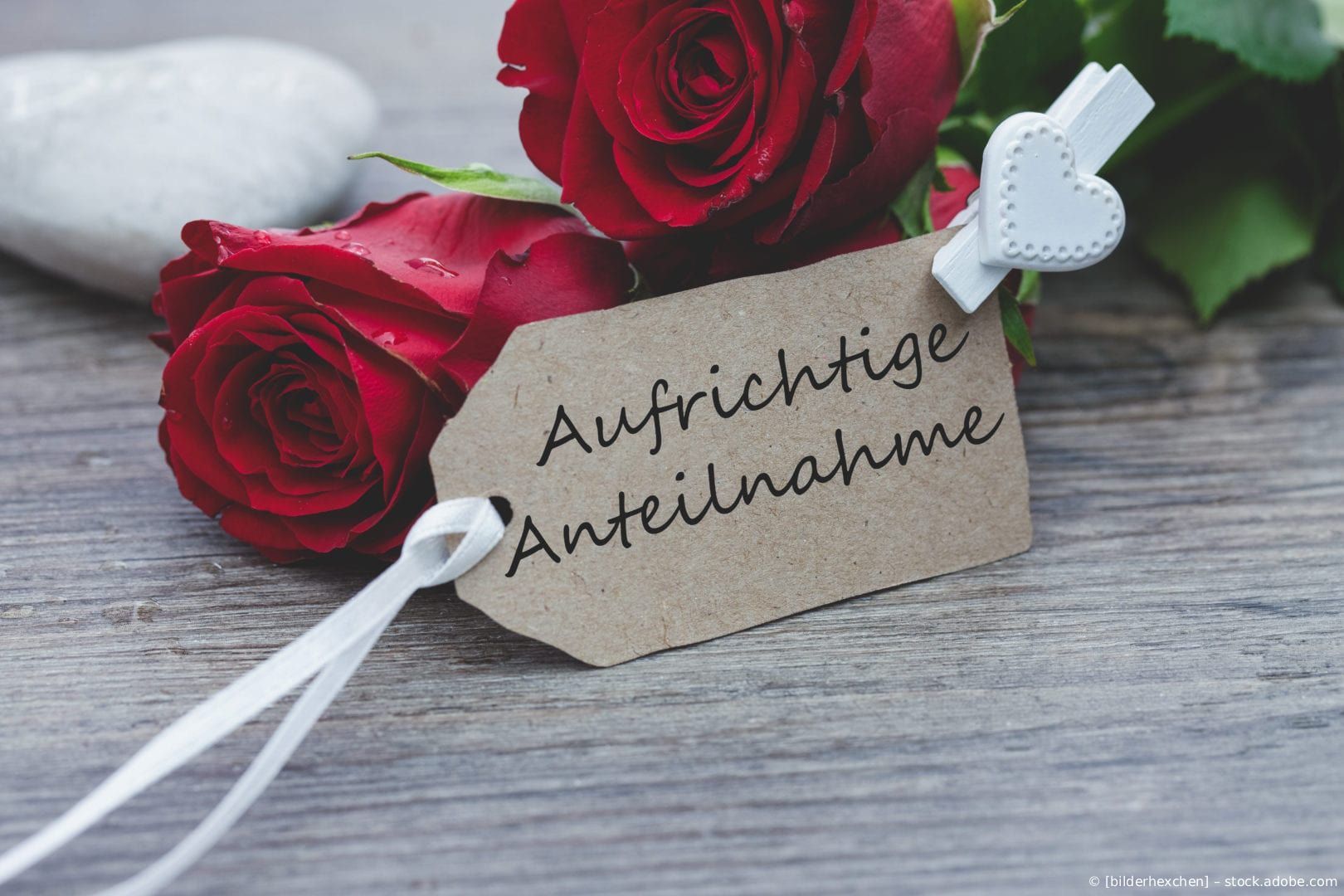Red roses and a white heart on a tag reading “Aufrichtige Anteilnahme” on a gray wooden surface