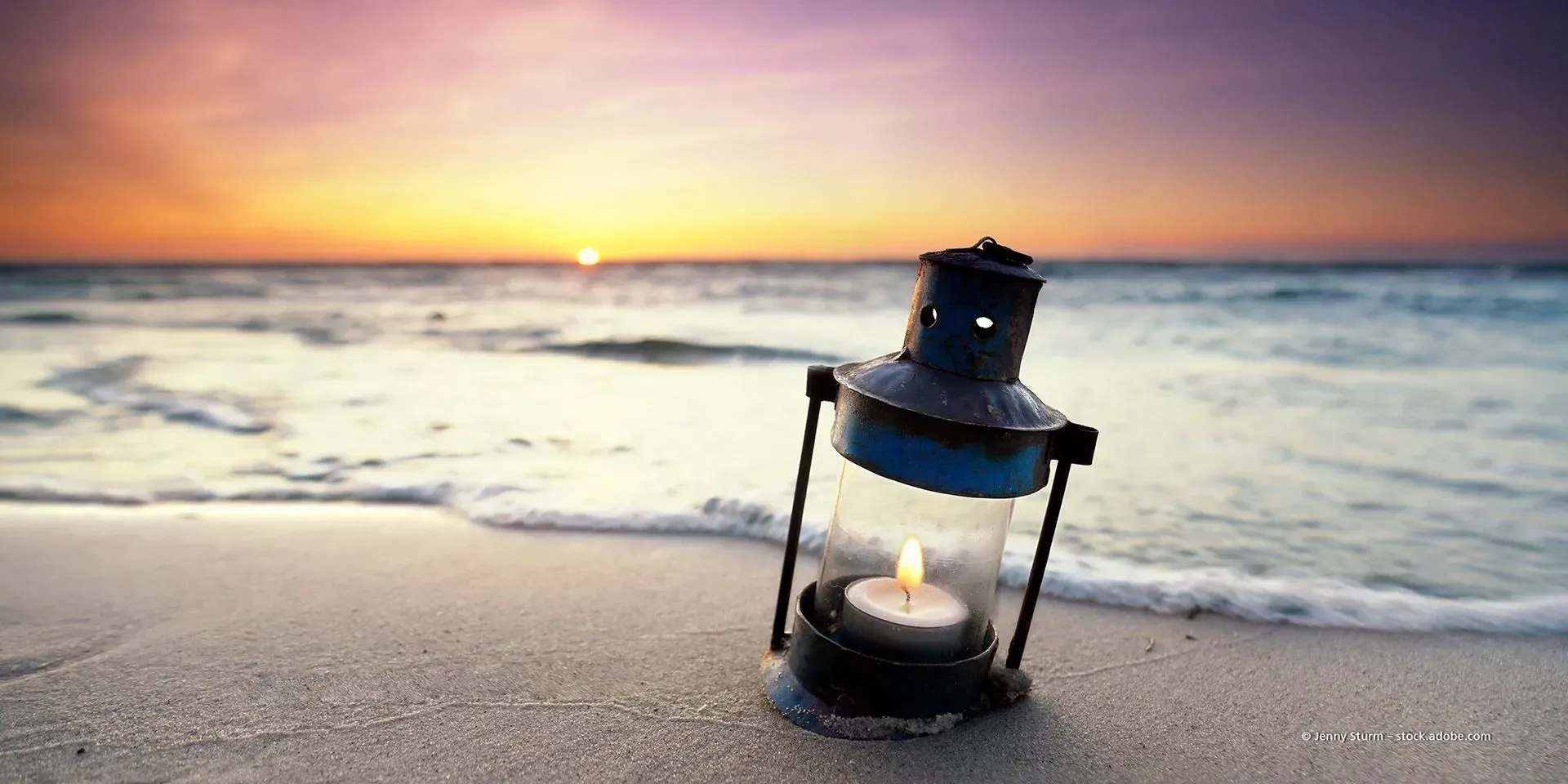 Eine beleuchtete Laterne an einem Sandstrand bei Sonnenuntergang, im Hintergrund die Wellen des Ozeans.