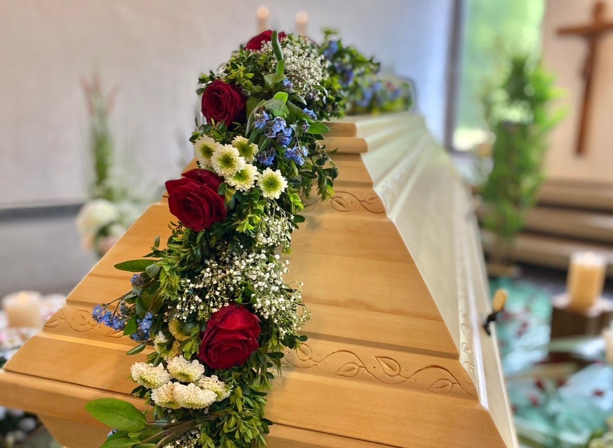 Blumenarrangement auf einem Holzsarg mit cremefarbenen und violetten Rosen bei einer Beerdigung