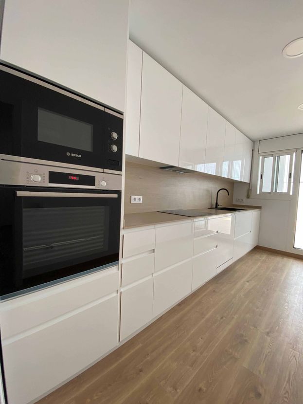 Cocina moderna blanca con horno empotrado, microondas y armarios. Suelo de madera clara y ventana a la derecha.