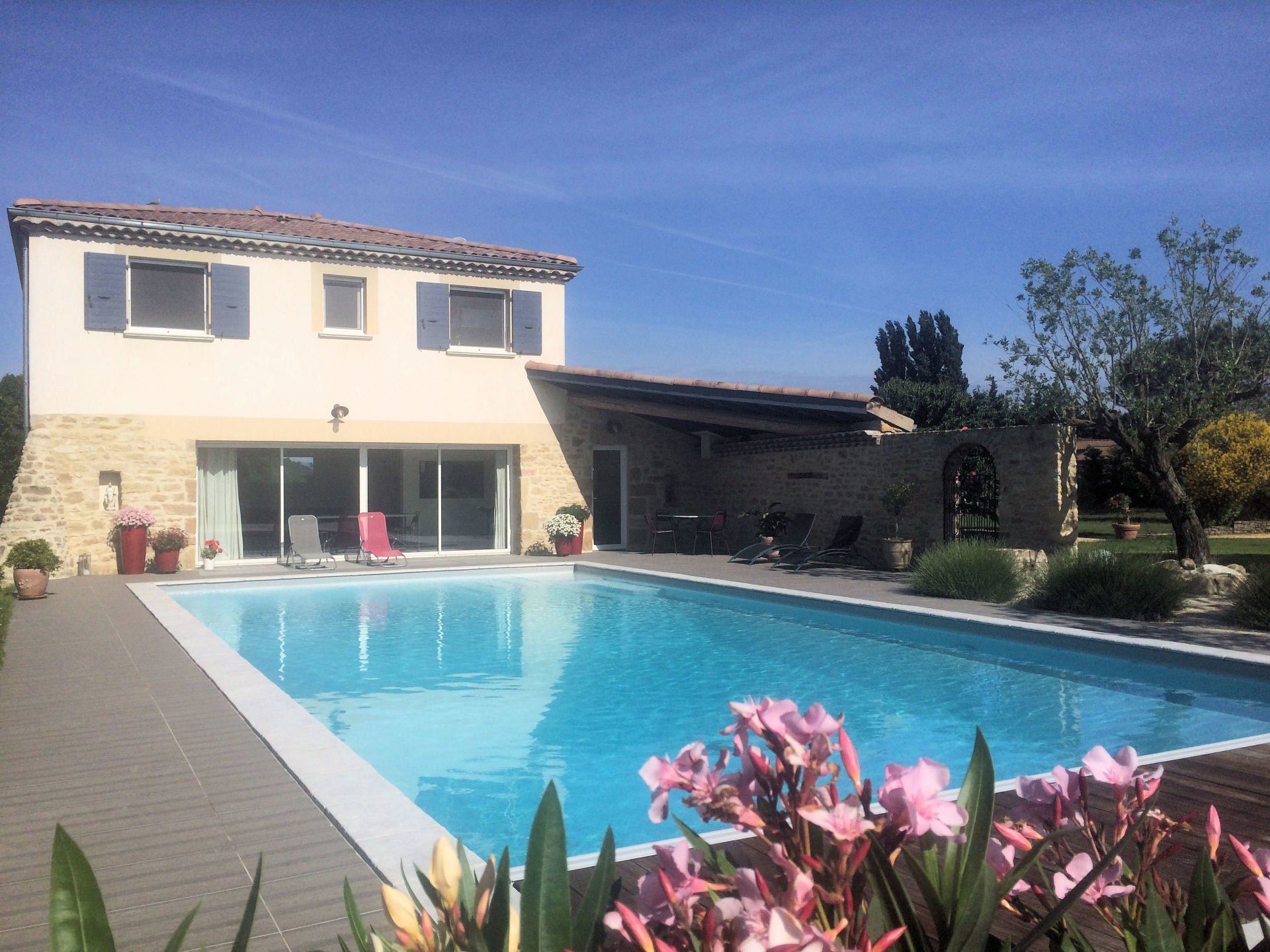 Une maison beige à deux étages avec piscine, terrasse et fleurs sous un ciel bleu.