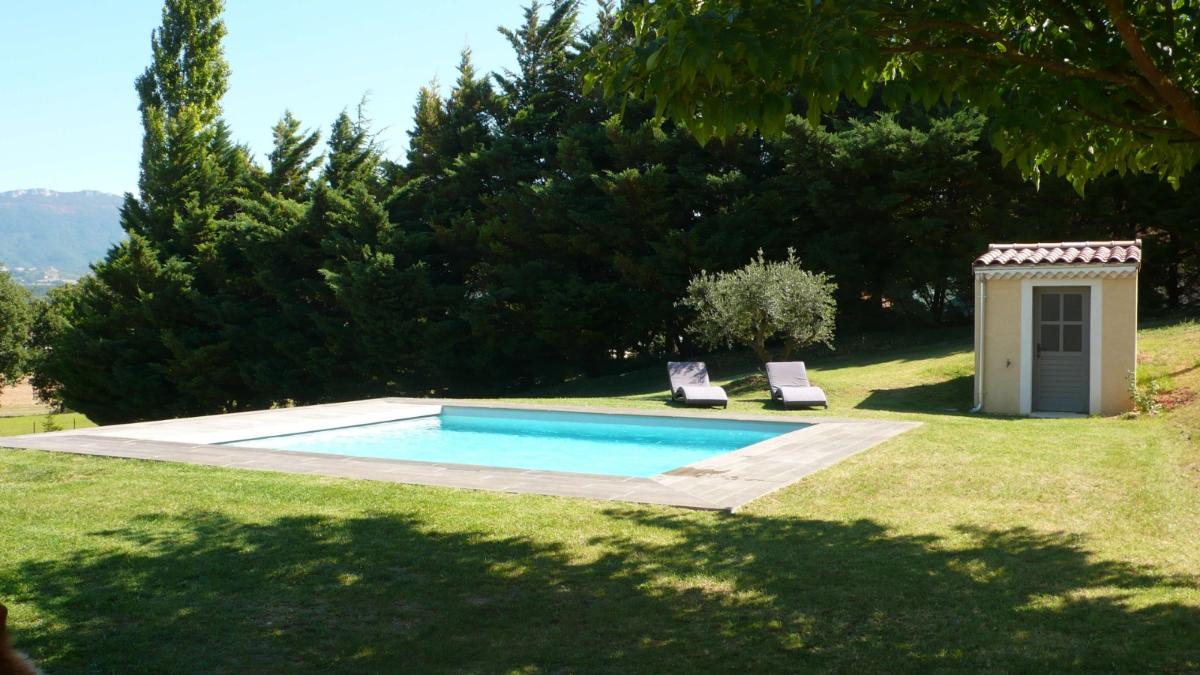 Piscine entourée de pelouse verte, d'arbres et d'un petit abri de jardin par une journée ensoleillée.