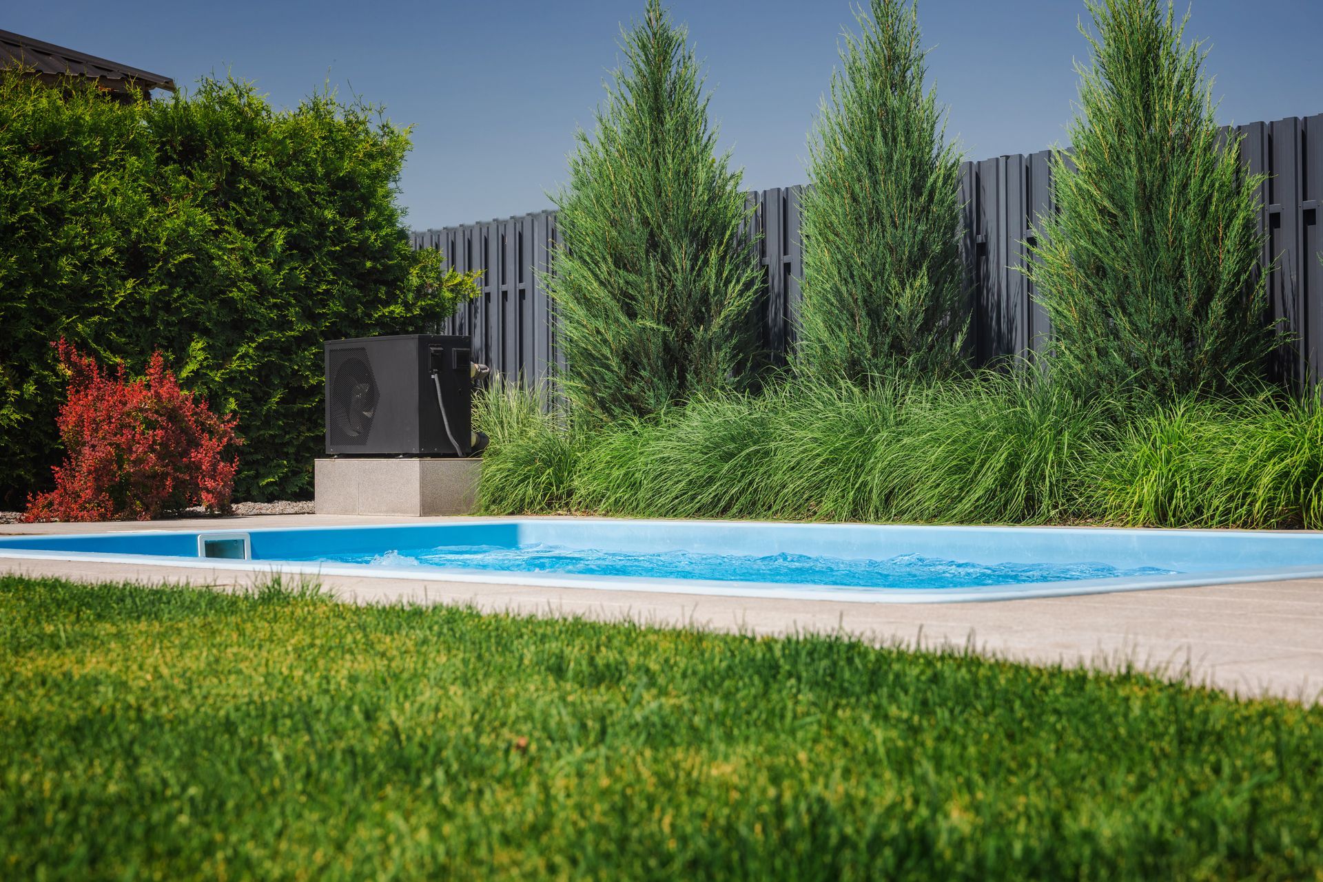 Piscine avec pompe à chaleur, pelouse verdoyante, eau bleue et clôture en arrière-plan.