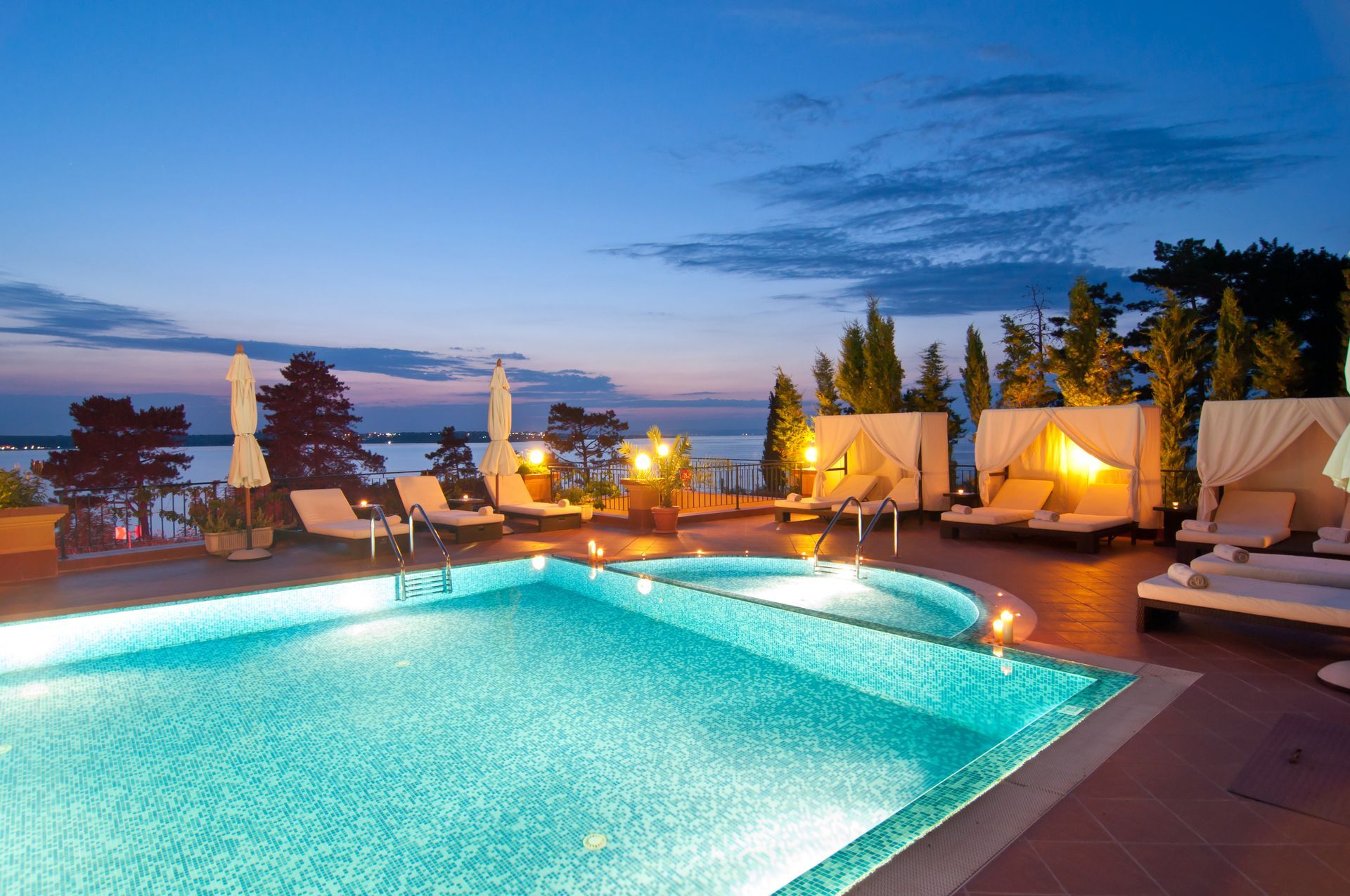 Piscine et espace détente avec vue sur l'eau au crépuscule ; éclairés par un éclairage d'ambiance.
