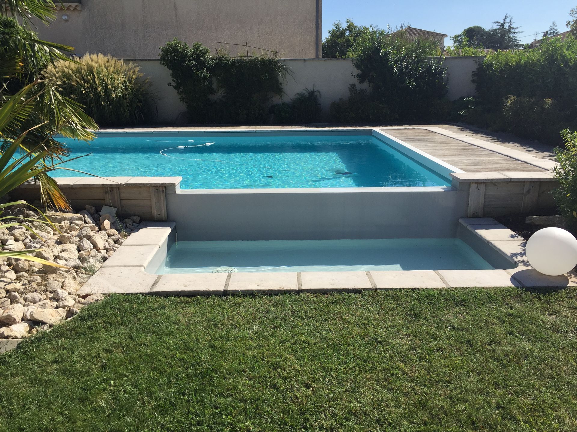 Piscine rectangulaire avec escalier, entourée de pelouse et d'aménagements paysagers par une journée ensoleillée.
