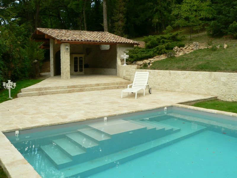 Piscine avec escalier, chaise longue et terrasse avec un petit bâtiment.