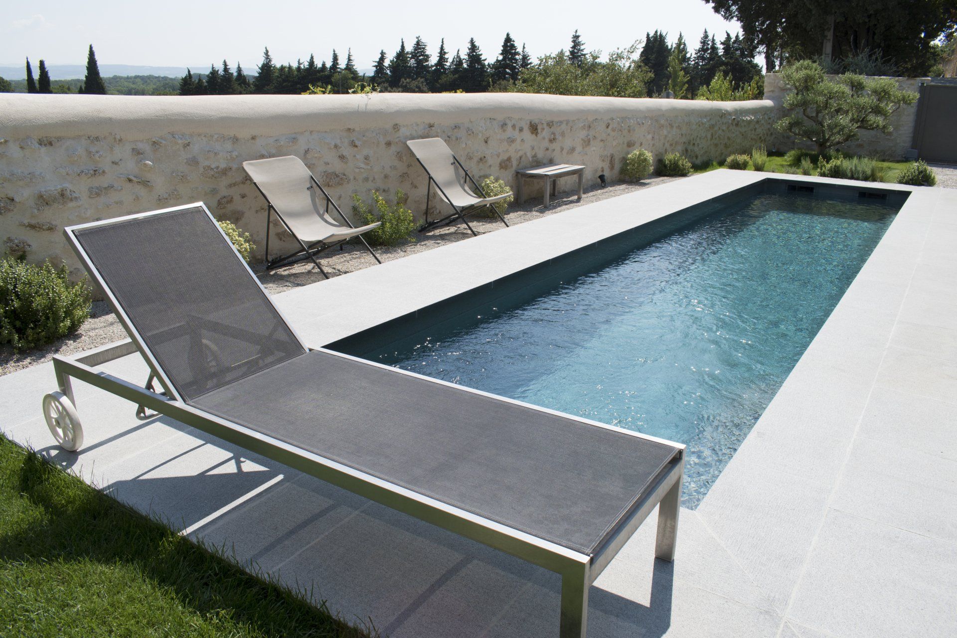 Chaise longue au bord d'une piscine rectangulaire aux eaux turquoise, adossée à un mur de pierre et à des arbres.