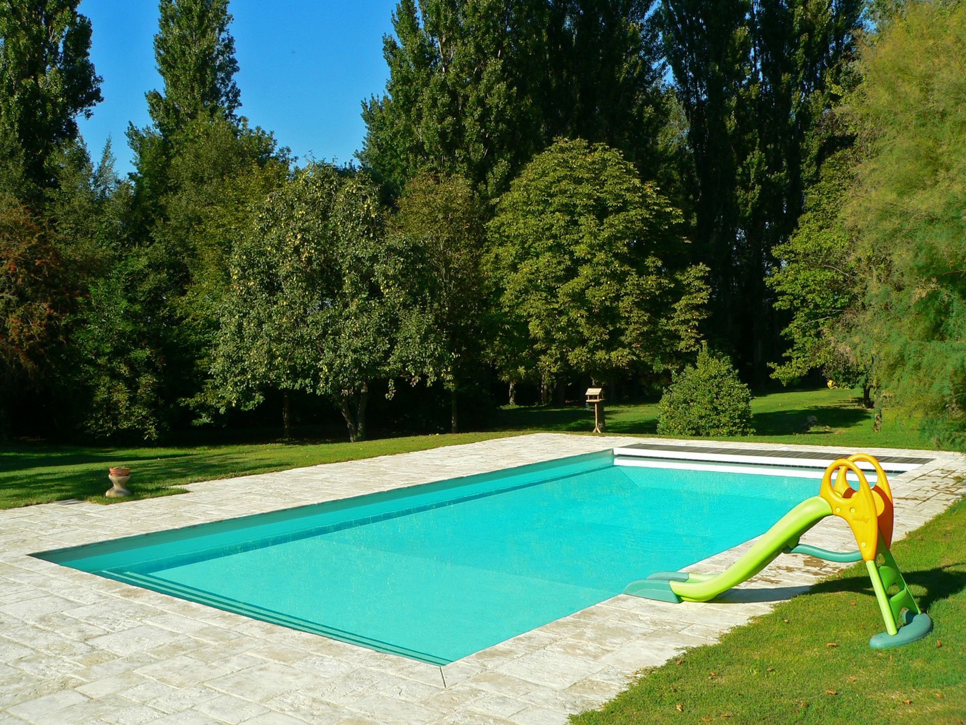 Piscine rectangulaire avec toboggan entourée de pelouse et d'arbres.
