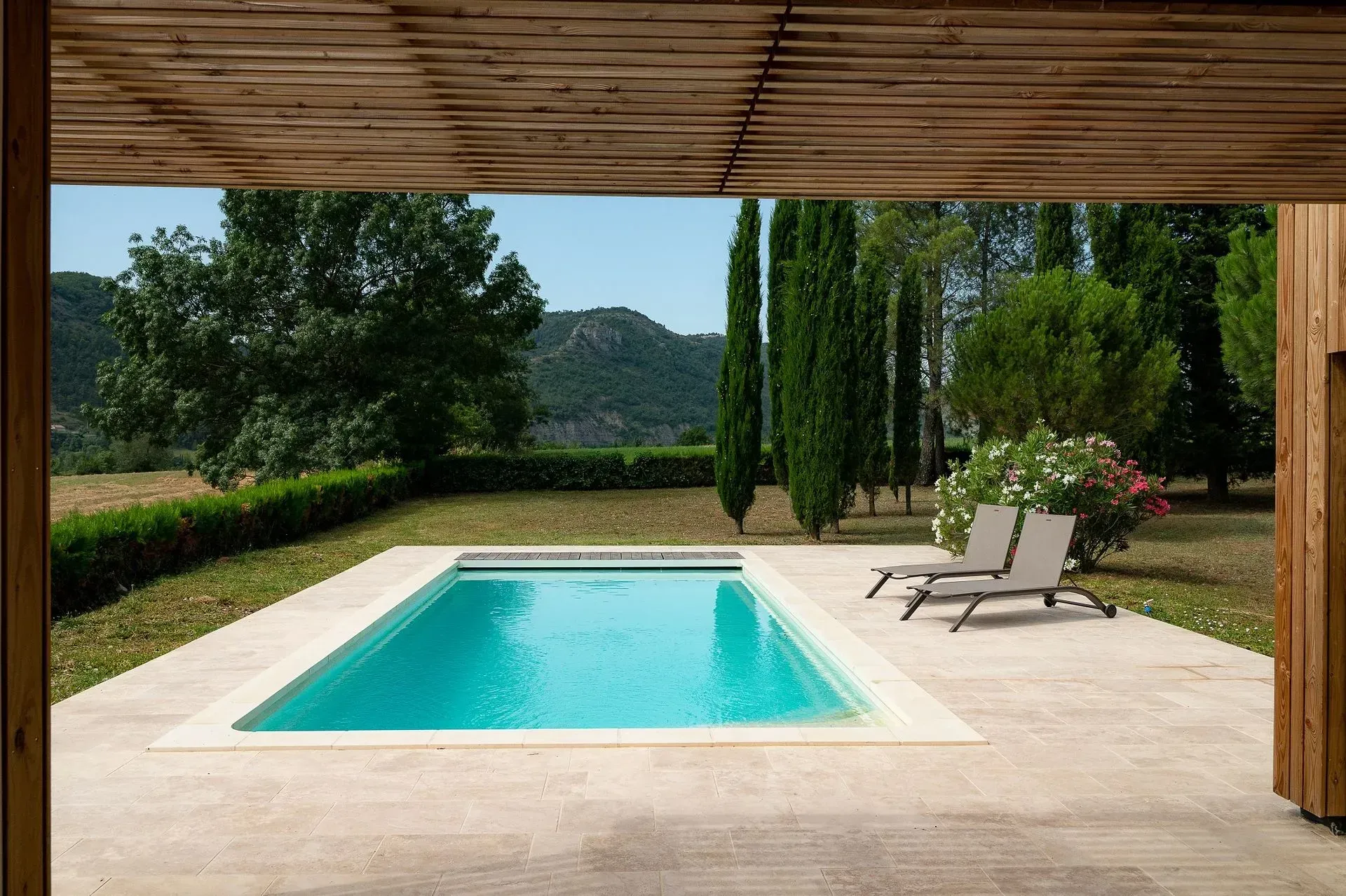 Piscine avec terrasse en bois, chaise longue, table avec chaises et arbres en arrière-plan.
