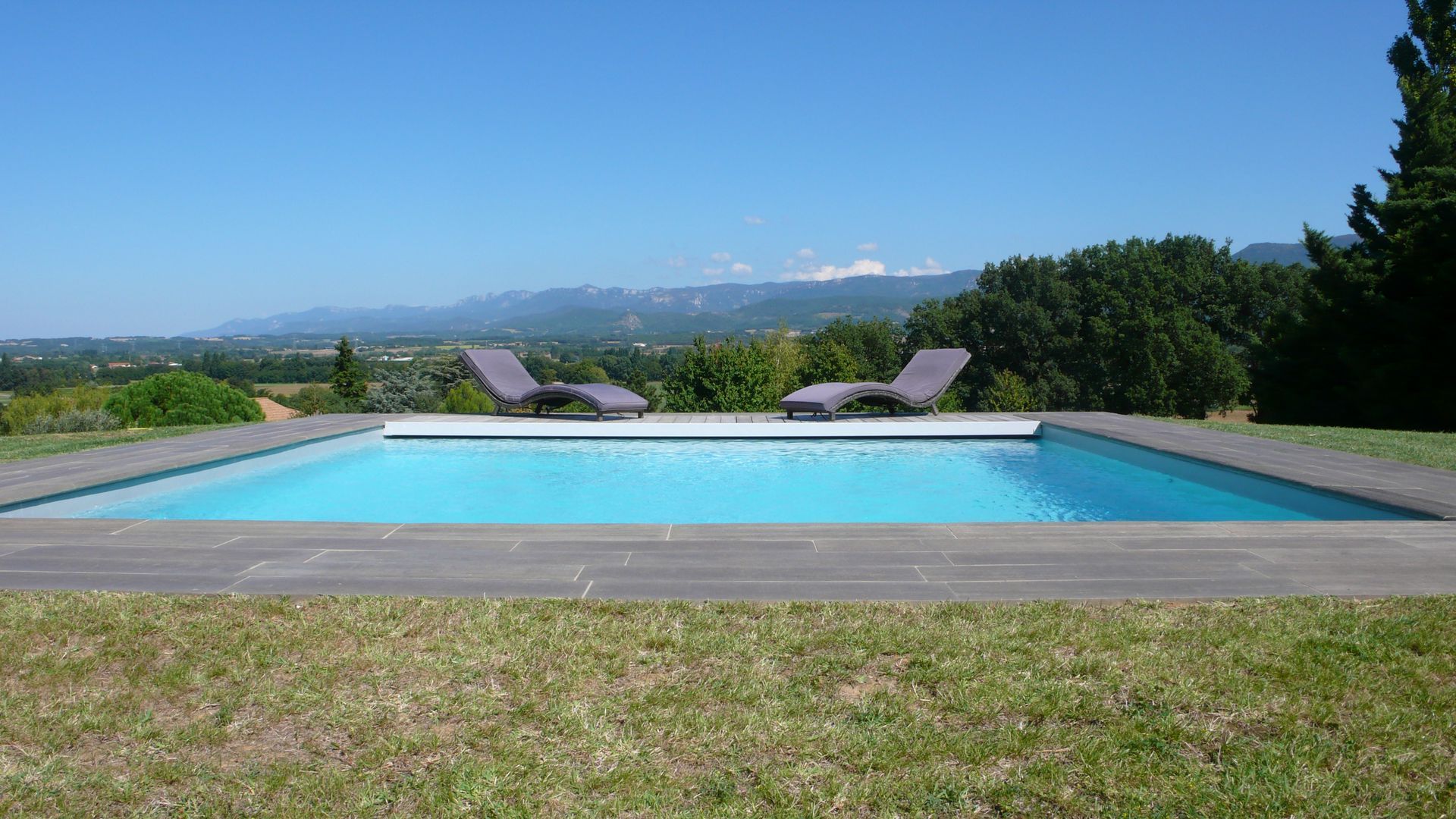 Piscine avec deux chaises longues, pelouse verte et vue sur les montagnes sous un ciel bleu.