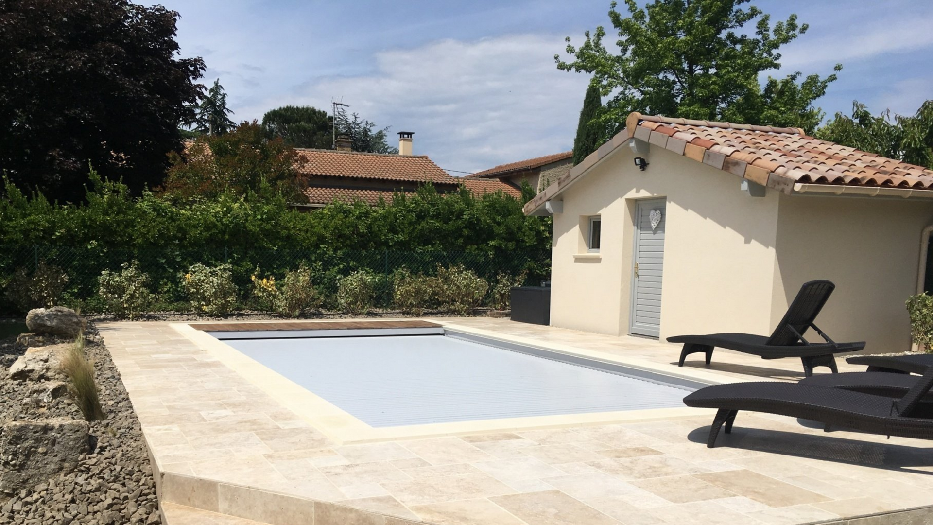 Piscine couverte, terrasse beige, petit bâtiment et chaises longues par une journée ensoleillée.
