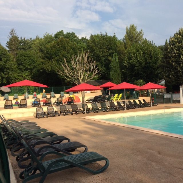 Scène au bord de la piscine avec parasols rouges, chaises longues et arbres verdoyants.