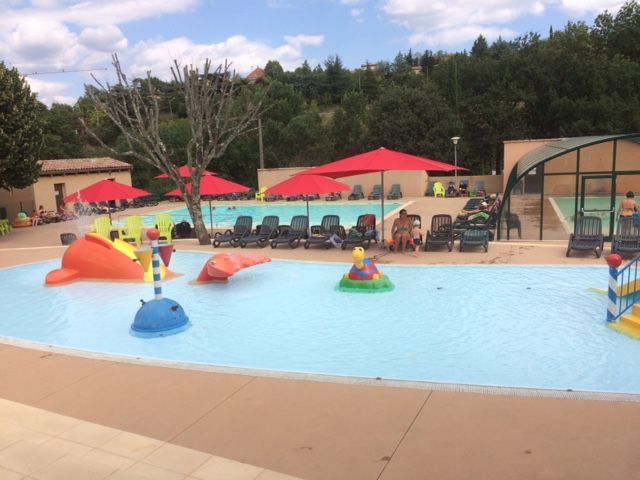 Piscine avec structures de jeux et chaises longues sous des parasols rouges par une journée ensoleillée.