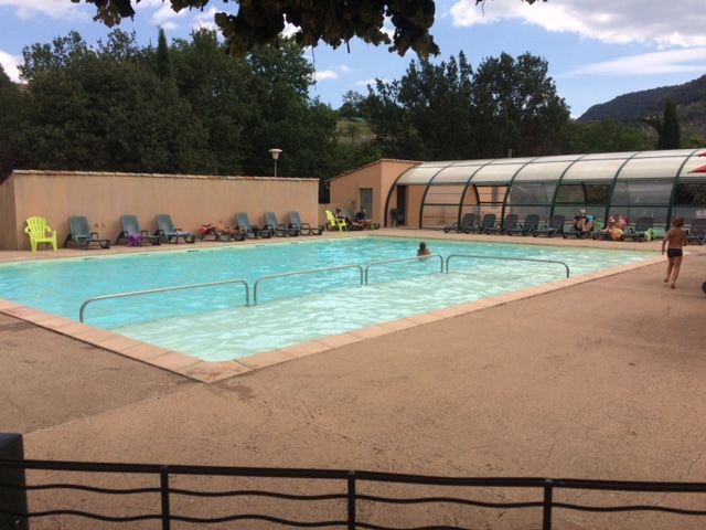 Piscine avec chaises longues, partiellement fermée, une personne nageant, arbres en arrière-plan.