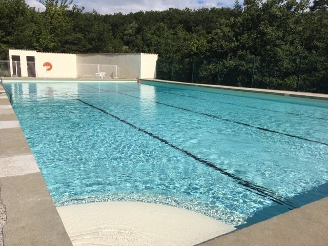 Piscine extérieure avec eau claire, ciel bleu et arbres. Entourage en béton, équipement de surveillance de baignade.