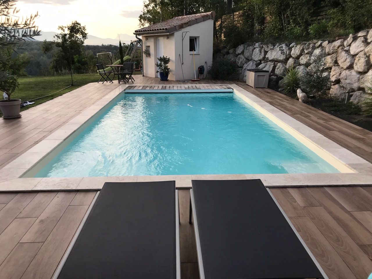 Piscine avec deux chaises longues, petit bâtiment et mur de pierre en arrière-plan.