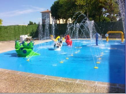 Une aire de jeux d'eau avec des fontaines, des figurines sur le thème du sport et un but jaune dans un parc.