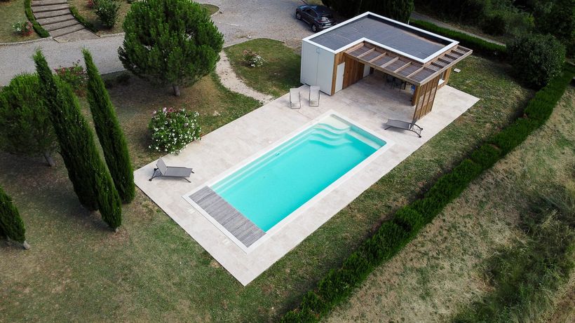 Une piscine rectangulaire aux eaux turquoise, entourée de béton, de pelouse et d'un petit bâtiment avec une pergola ombragée.