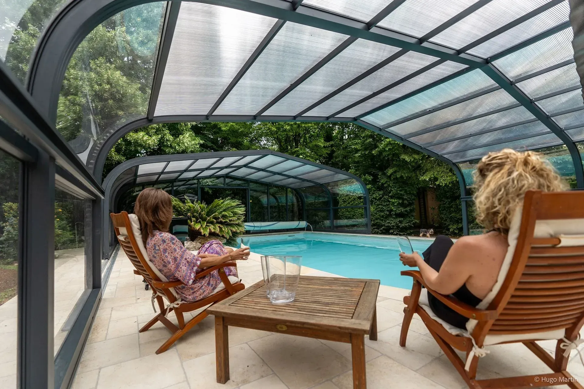 Deux personnes se détendent sur des chaises longues au bord d'une piscine couverte d'une structure en verre.