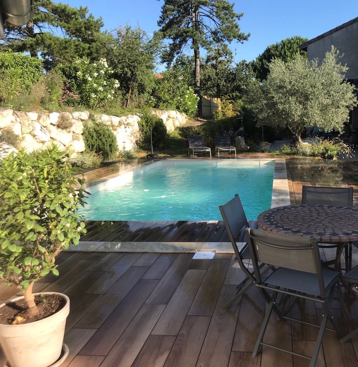 Une piscine dans un jardin avec terrasse en bois, table et chaises, entourée d'arbres et de verdure.