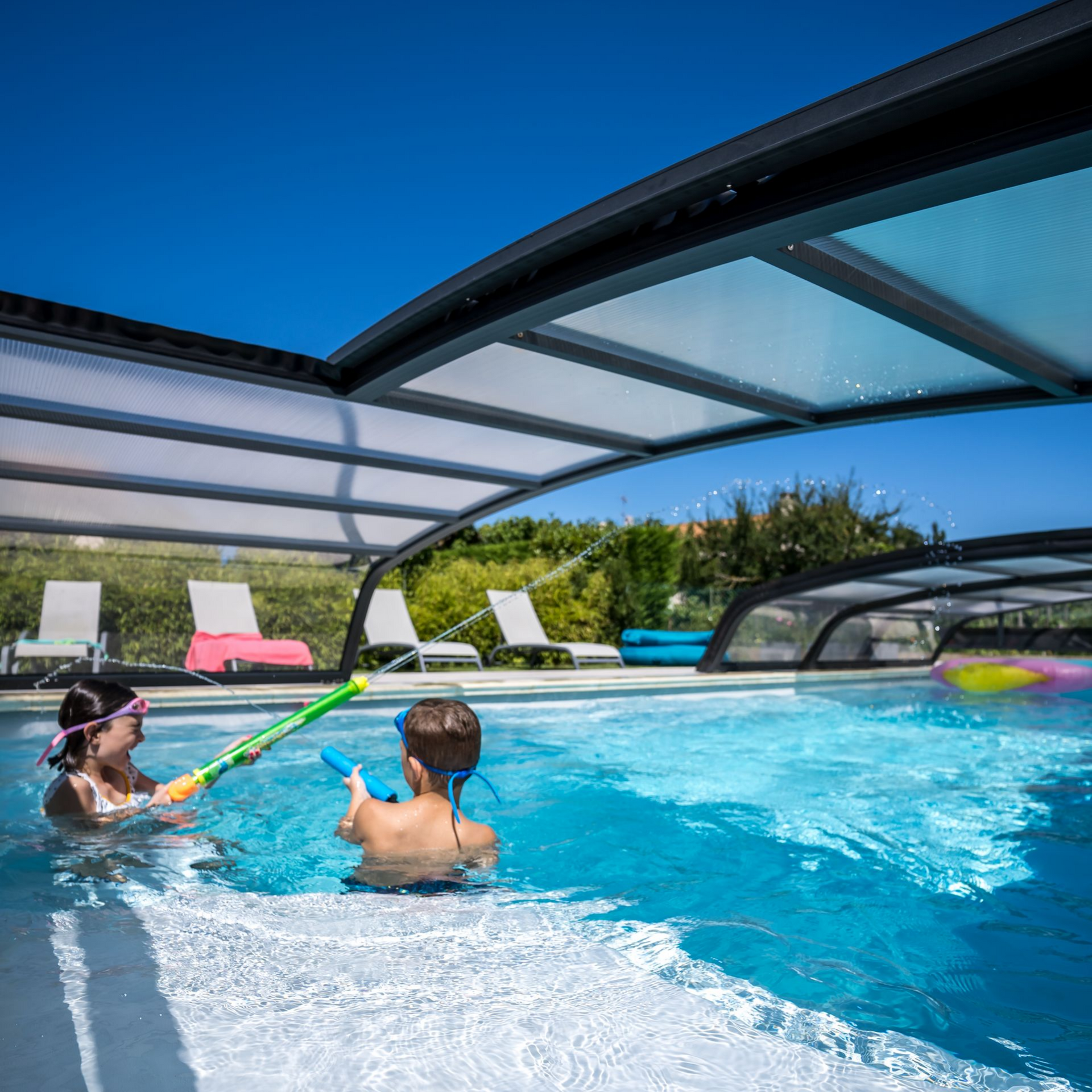 Une piscine rectangulaire entourée d'une couverture transparente, située dans un jardin avec une maison en pierre.