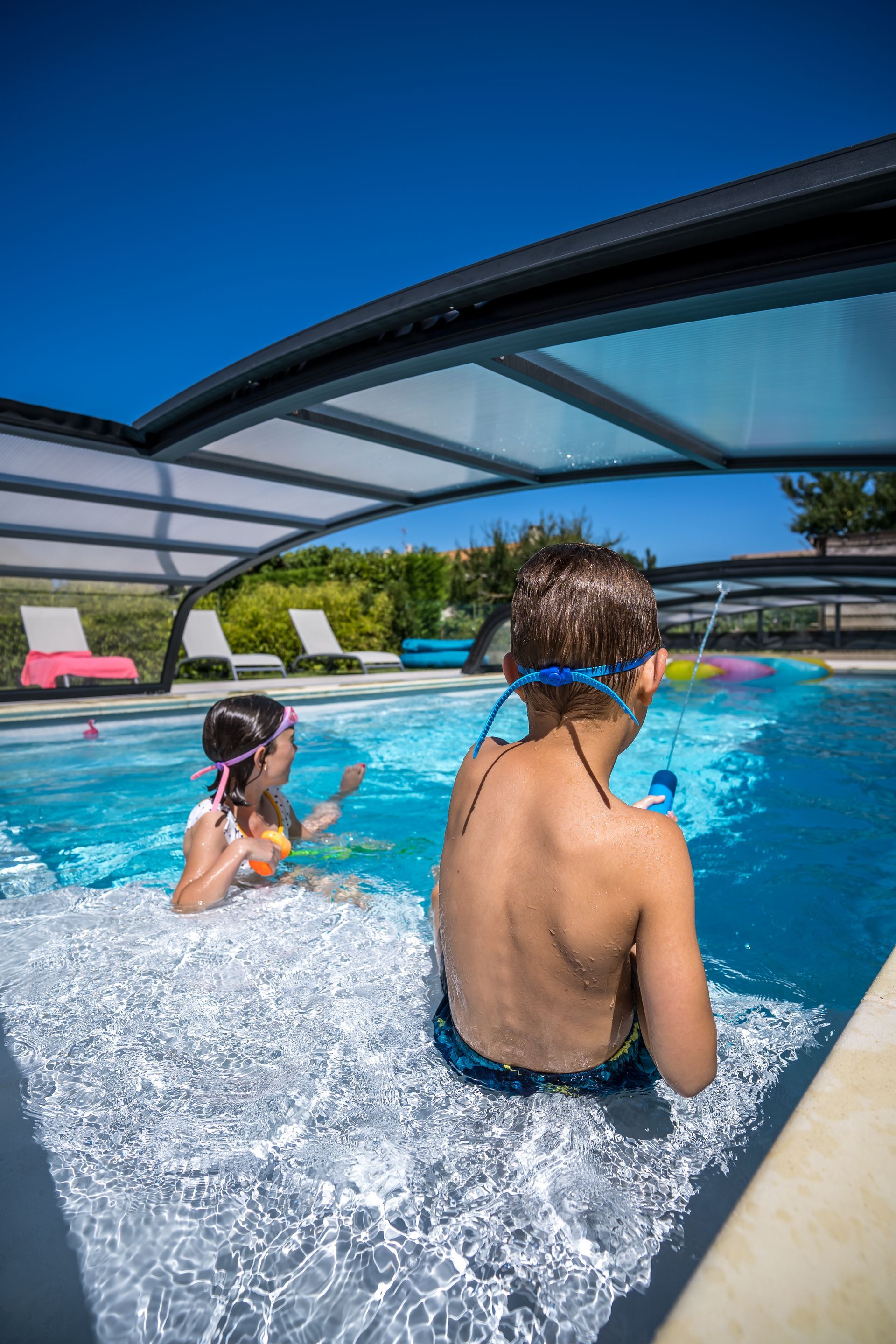 Piscine rectangulaire turquoise encadrée de pierre beige, vue depuis l'intérieur d'une structure en bois avec vue sur les arbres et les montagnes.