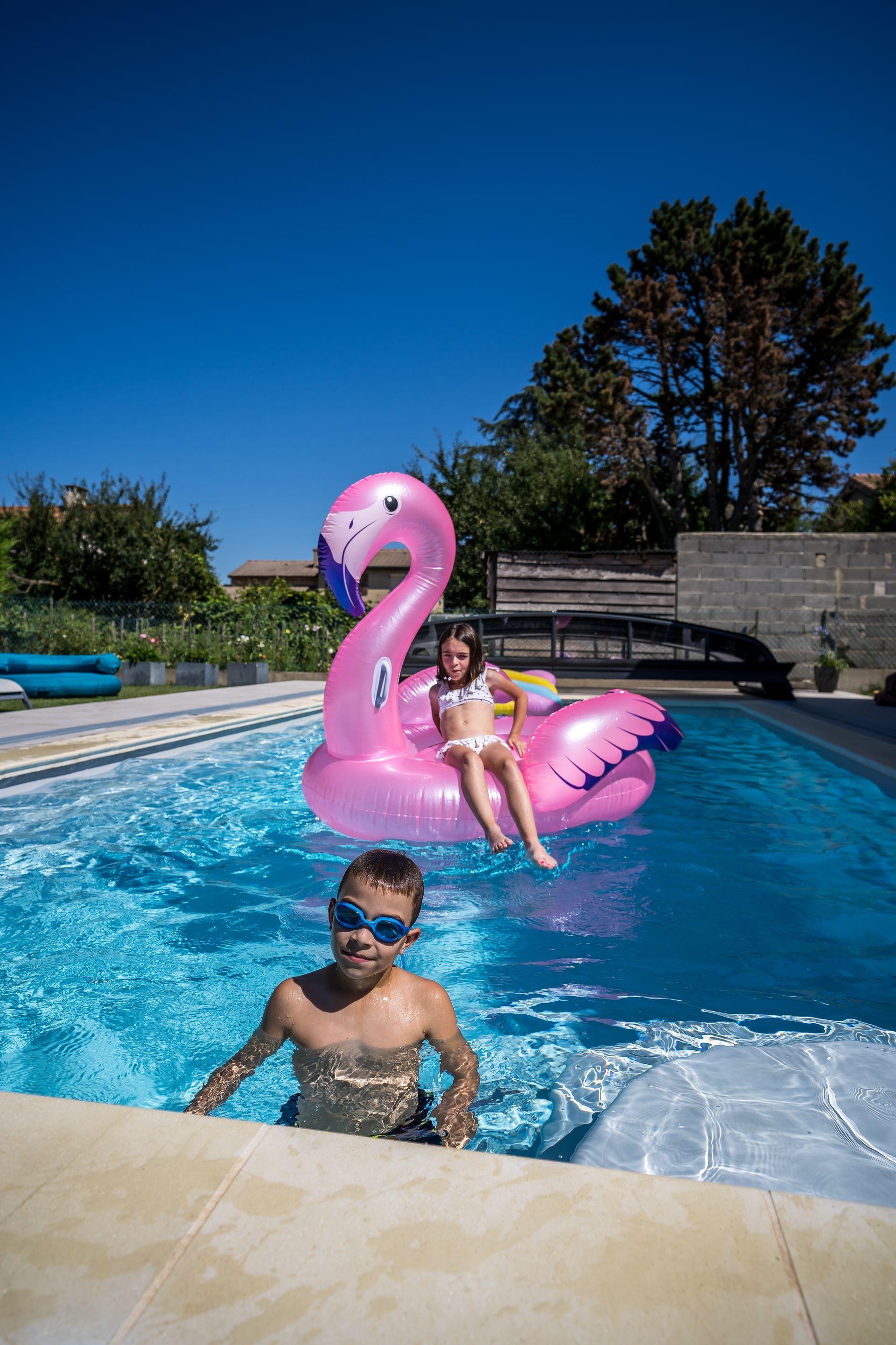 Un garçon dans une piscine avec une fille sur un flamant rose gonflable. Ciel bleu.